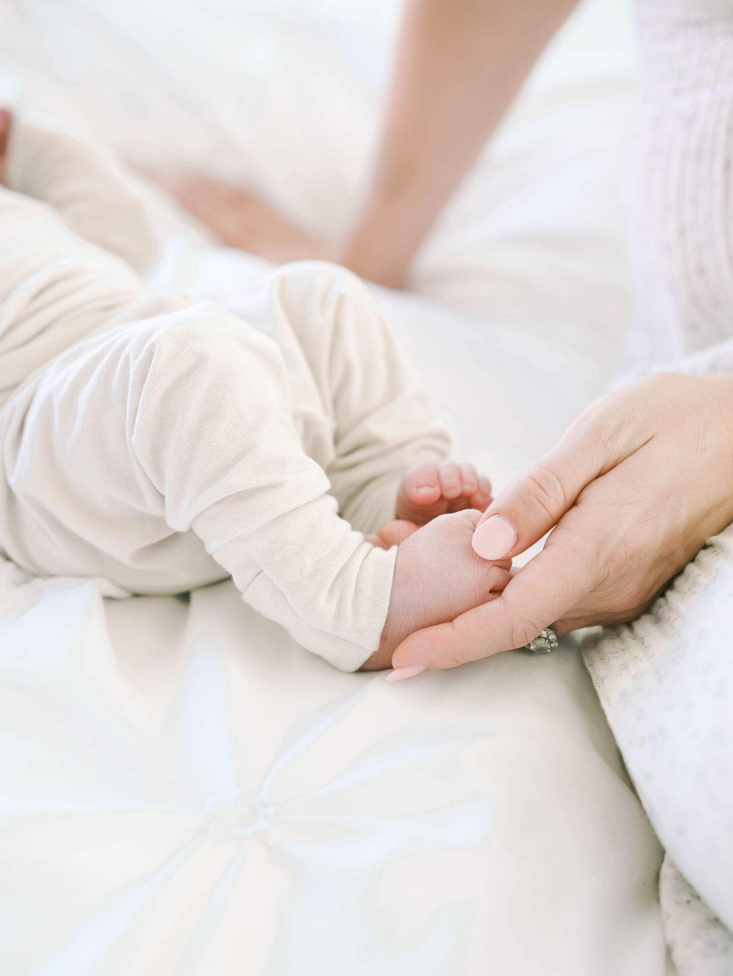 Mother gently holding newborn baby's hand while infant rests on white bedding.