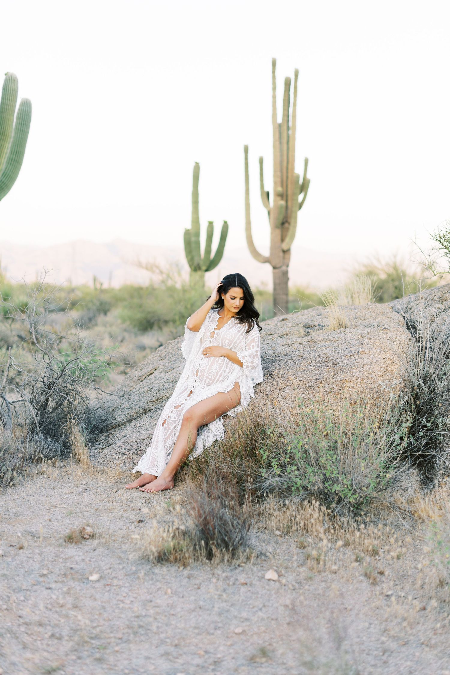 Pregnant brunette in white lace maxi dress sitting on desert rocks near saguaro cacti.