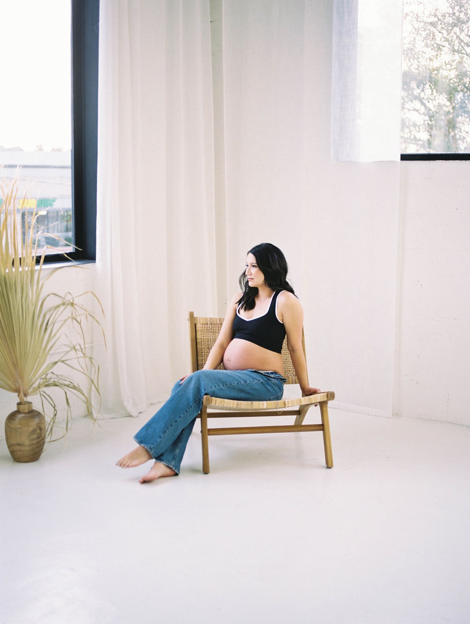 A pregnant woman in a black tank top and jeans sits relaxed in a rattan chair in a bright studio.