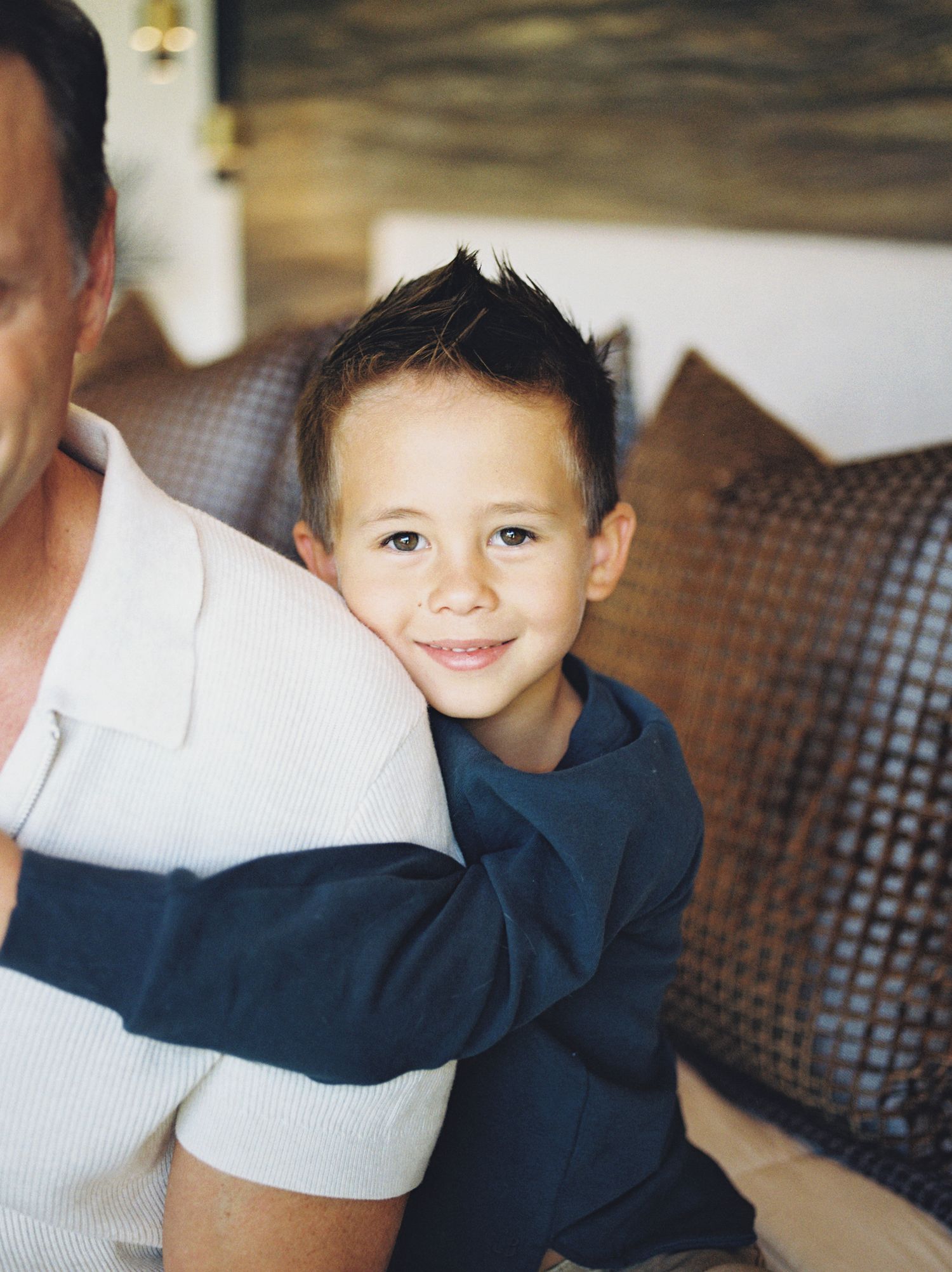 Smiling young boy with dark hair hugs an adult while sitting on a couch with brown pillows.