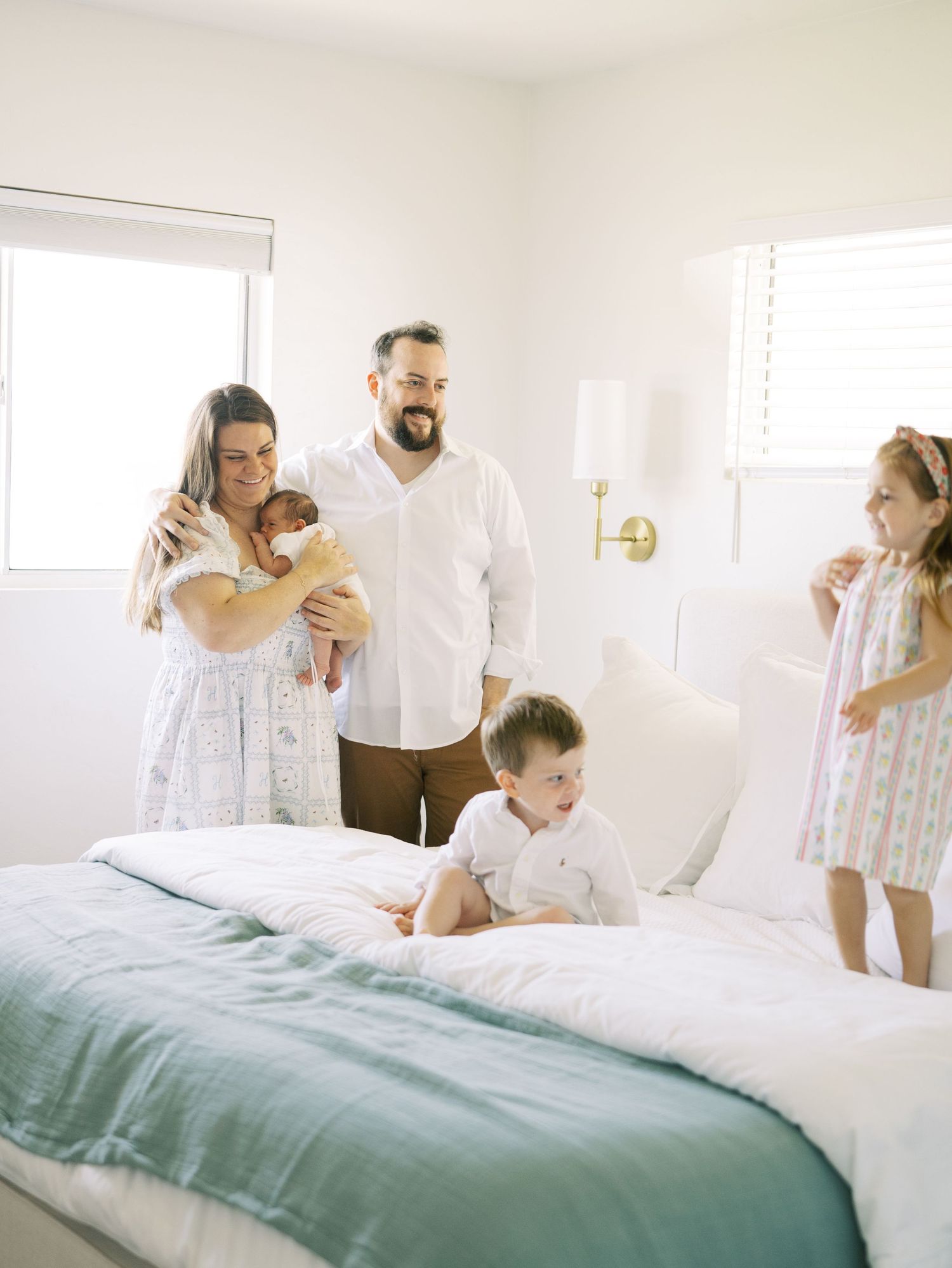 Family of four in bright white bedroom with parents holding newborn while children play on bed.