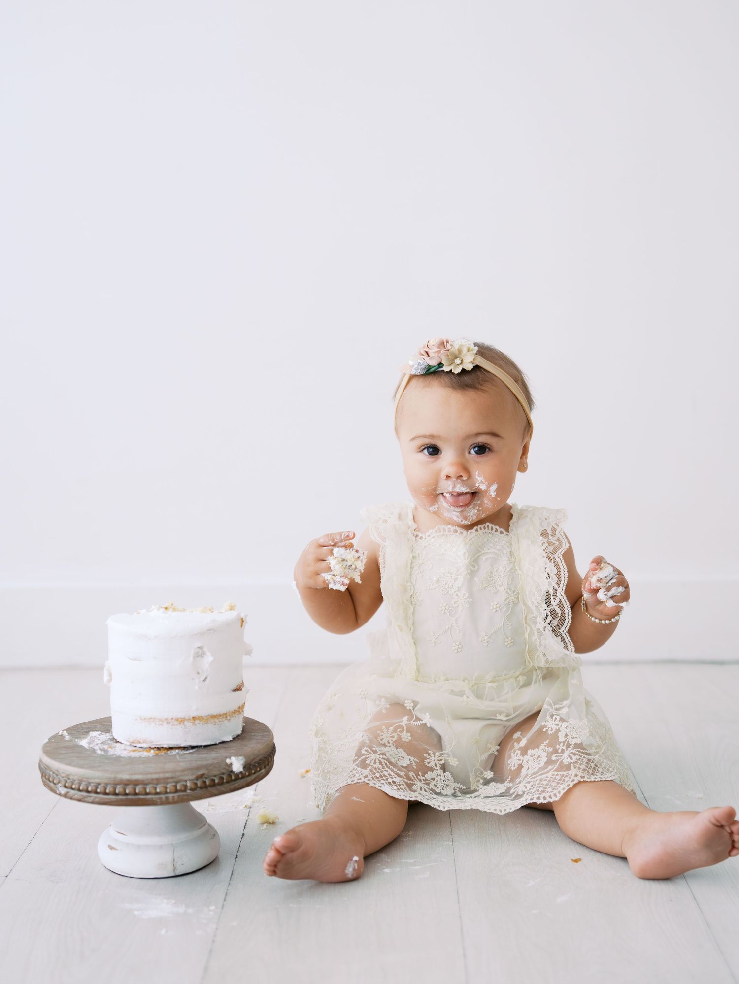 Smiling baby girl in lace dress with frosting on face next to smashed first birthday cake.