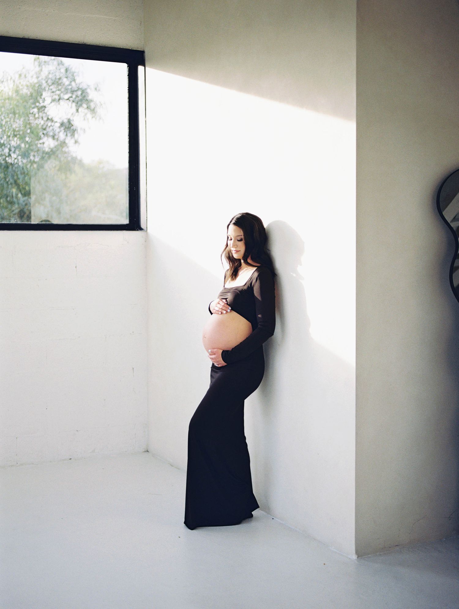 Pregnant woman in black gown leaning against white wall cradling baby bump in sunlight.