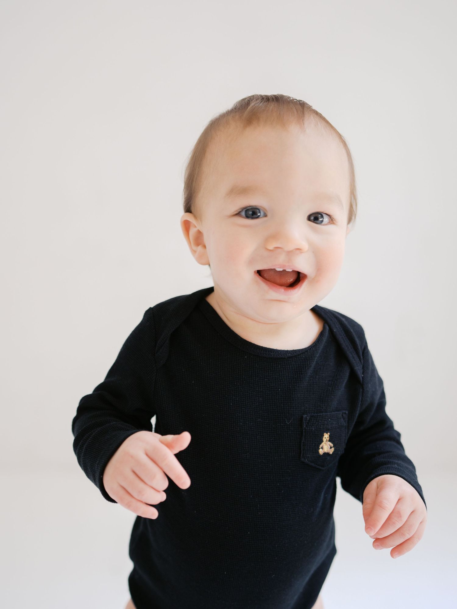 Happy smiling baby boy in black long sleeve onesie with gold bear patch on white background.