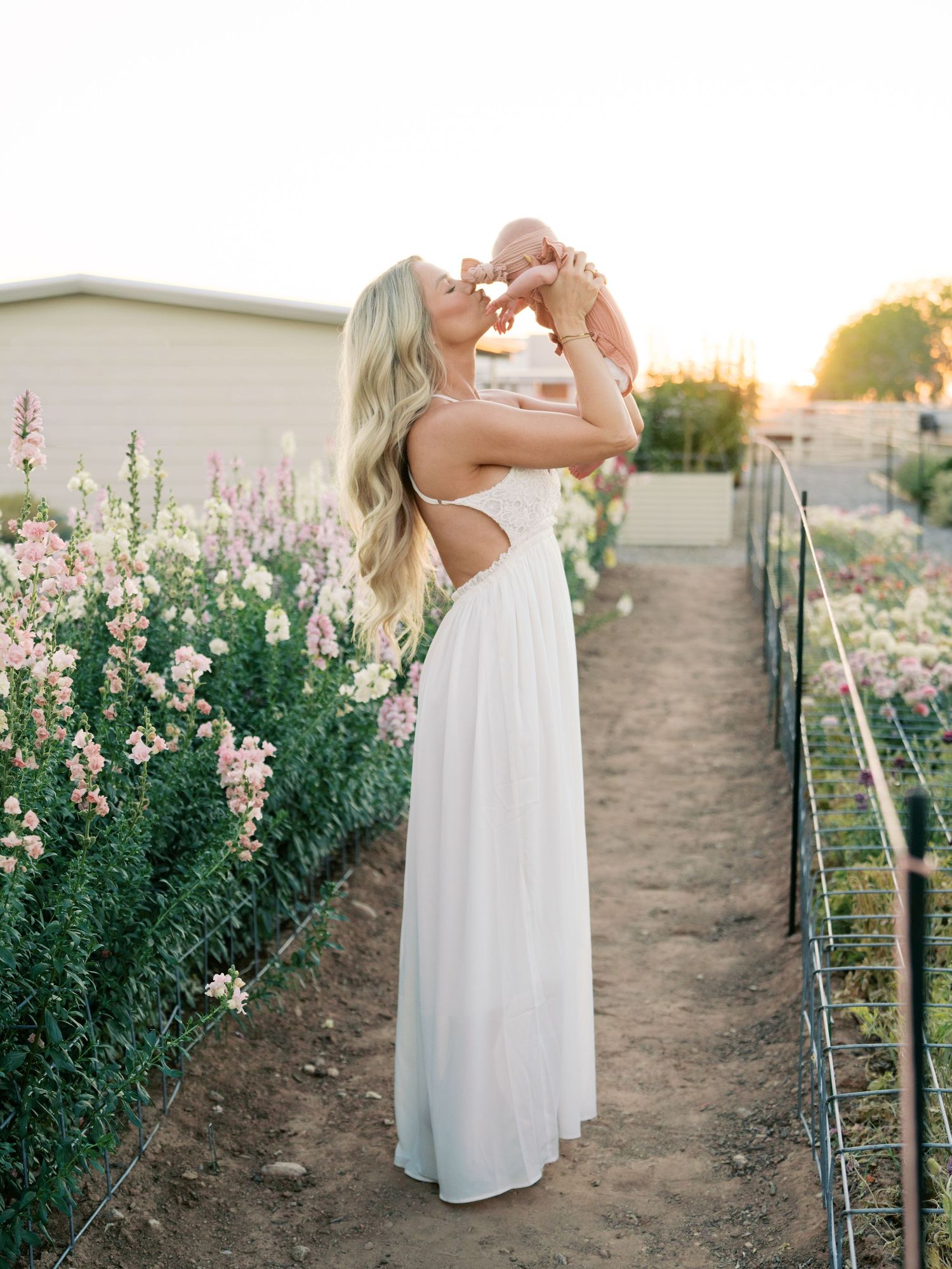 Blonde mother in white backless dress lifting smiling newborn baby at golden hour in flower garden.