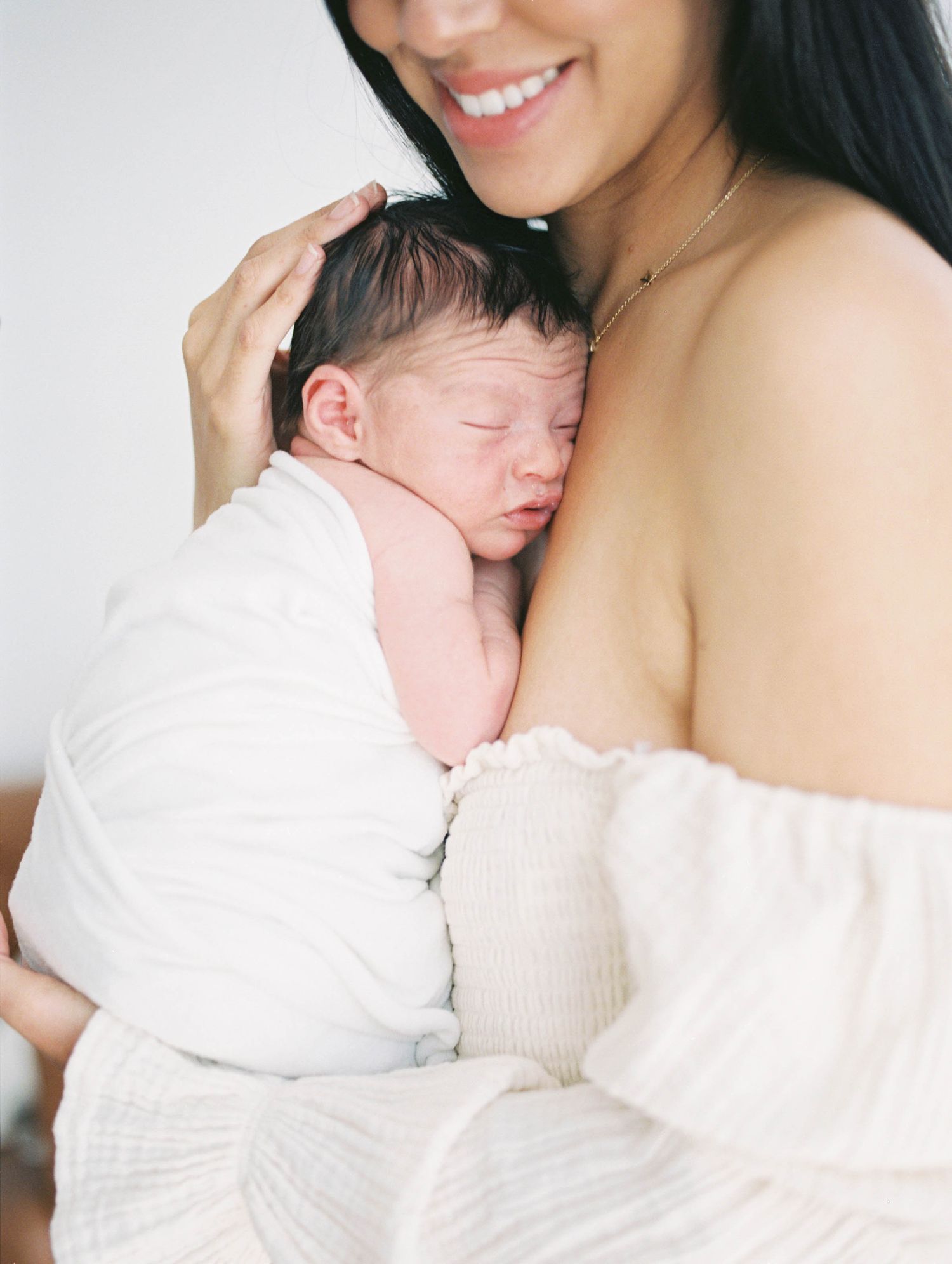 Close-up of smiling dark-haired mother in white off-shoulder top tenderly holding sleeping newborn.