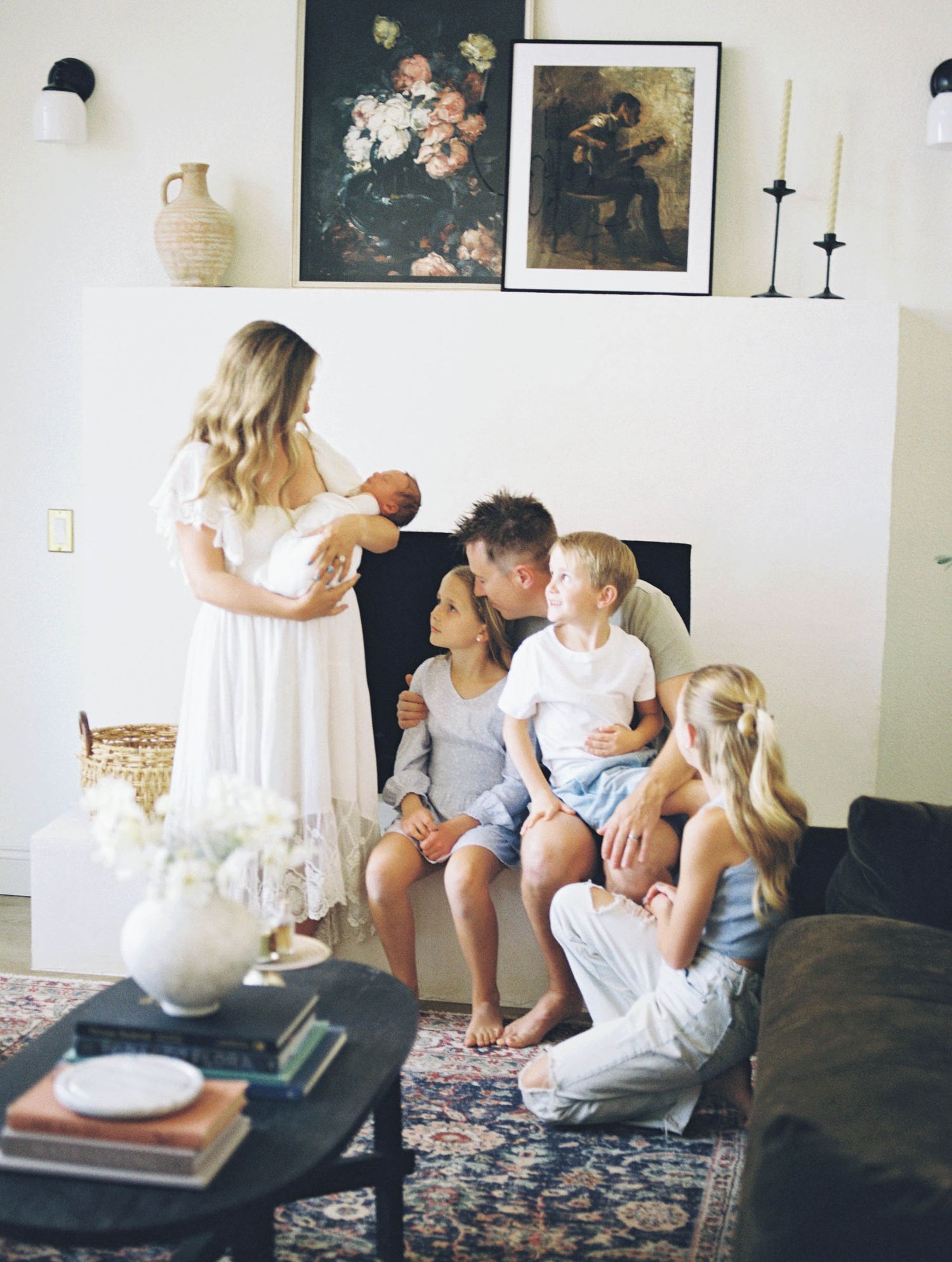 Blonde mother holding newborn while father kisses older child surrounded by three kids in living room.