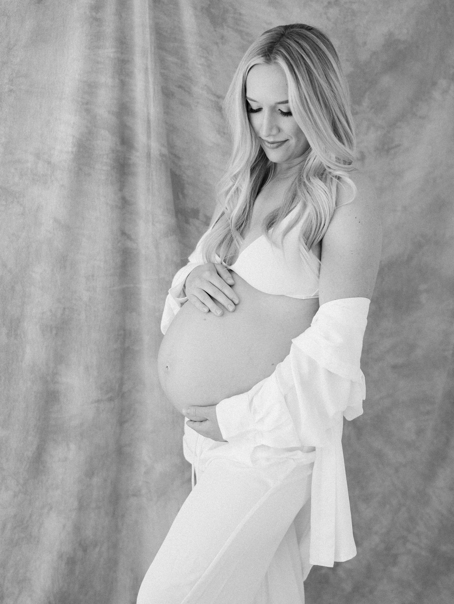 A black and white portrait of a pregnant woman in a white top gently cradling her baby bump in a studio.