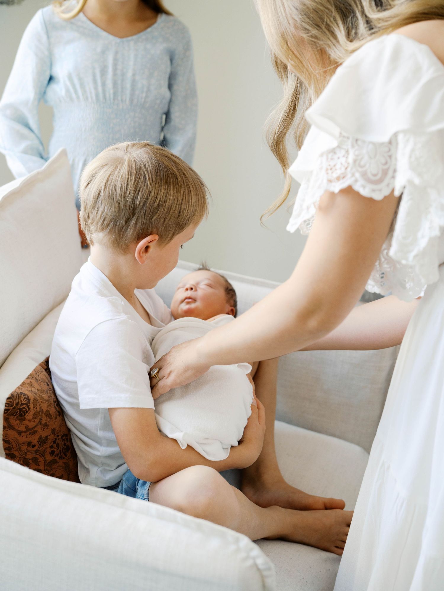 Young blonde boy carefully holding swaddled newborn sibling while seated in cream nursery chair.