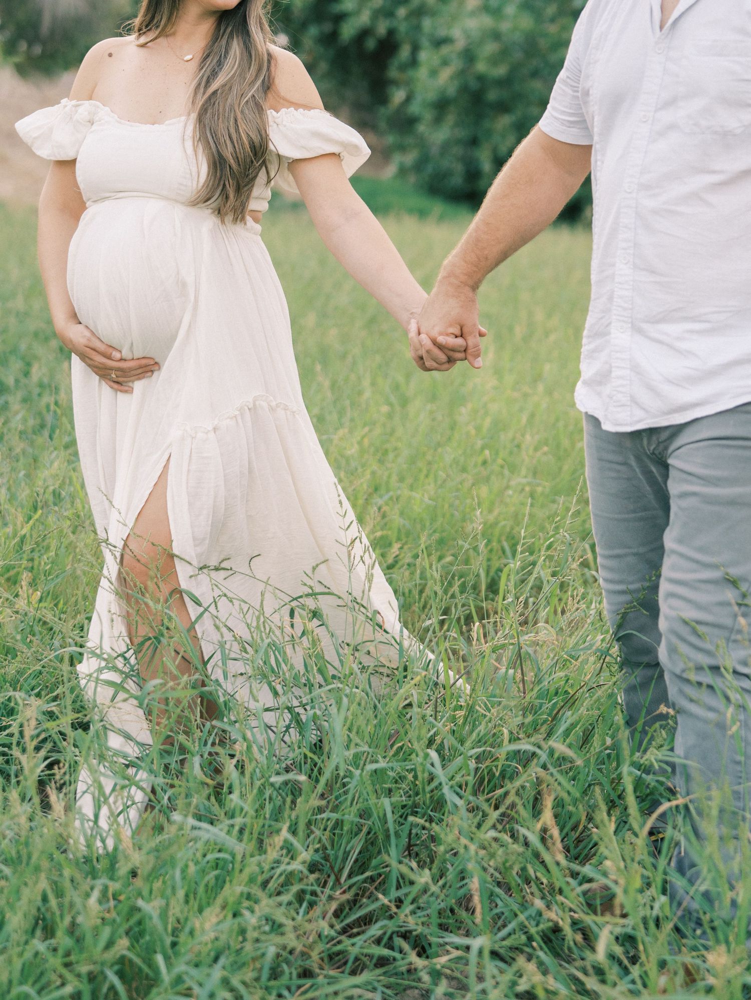 Pregnant woman in white off-shoulder dress holding hands with partner while walking through tall grass.