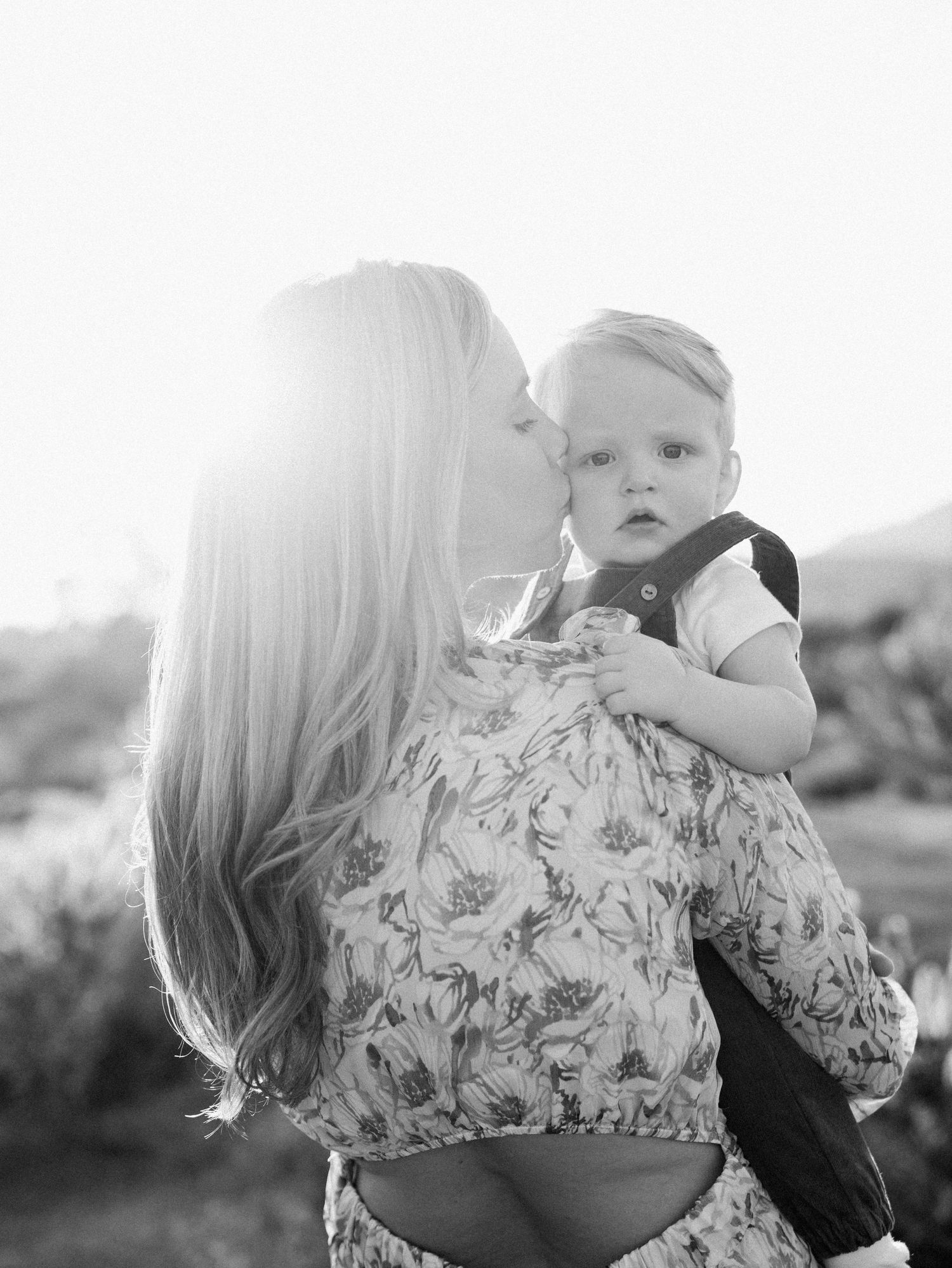 Black and white image of blonde mother kissing baby boy in carrier in bright sunlight.