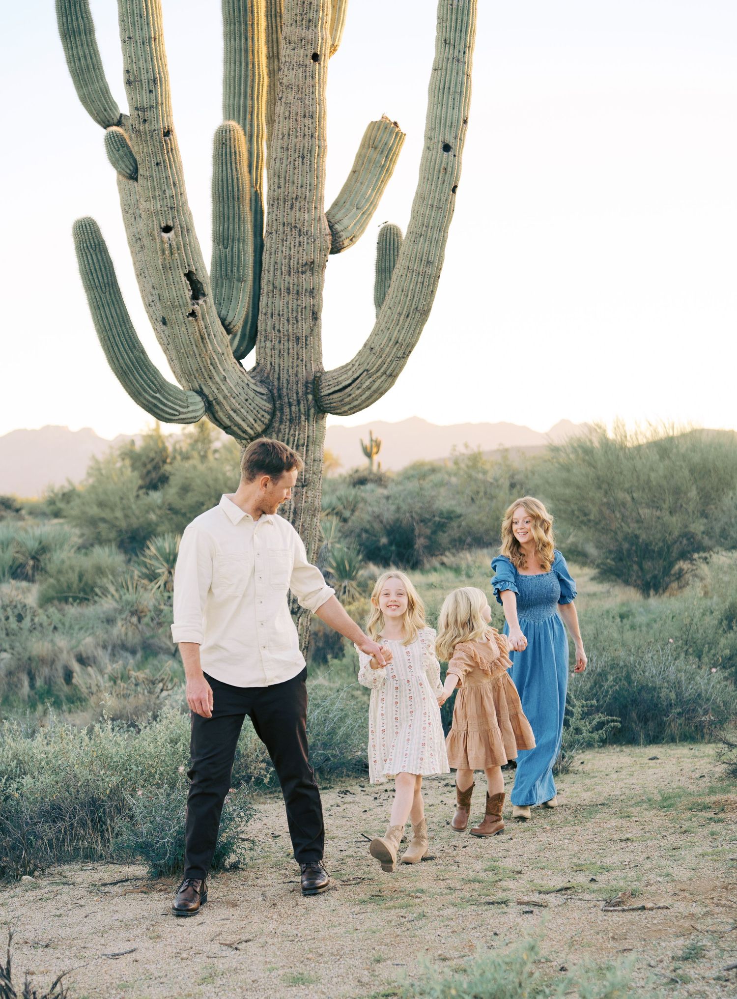 Family of four walking hand in hand near giant saguaro cactus at sunset.