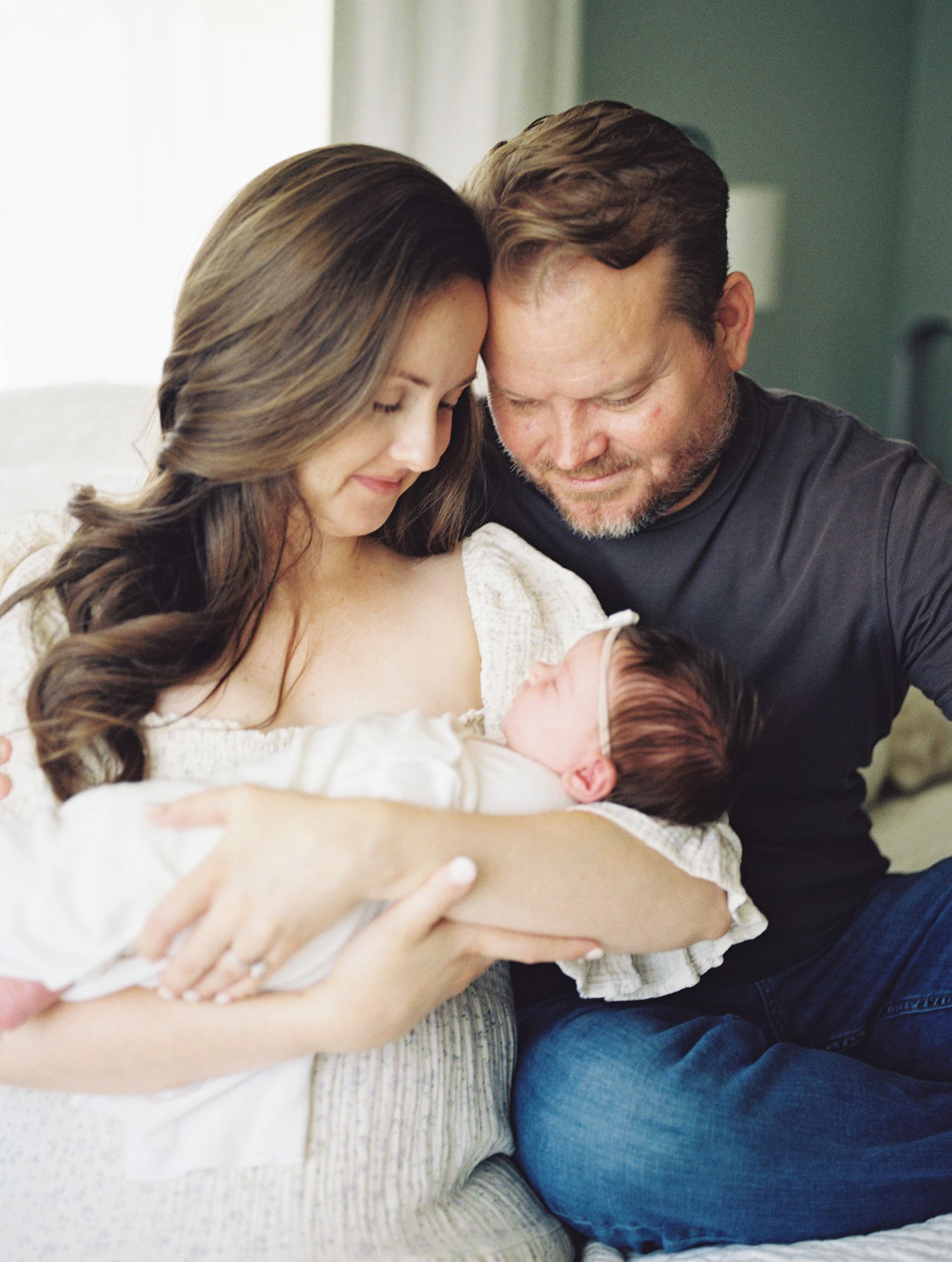 Loving parents with foreheads touching gently cradle sleeping newborn baby girl in soft white nursery.