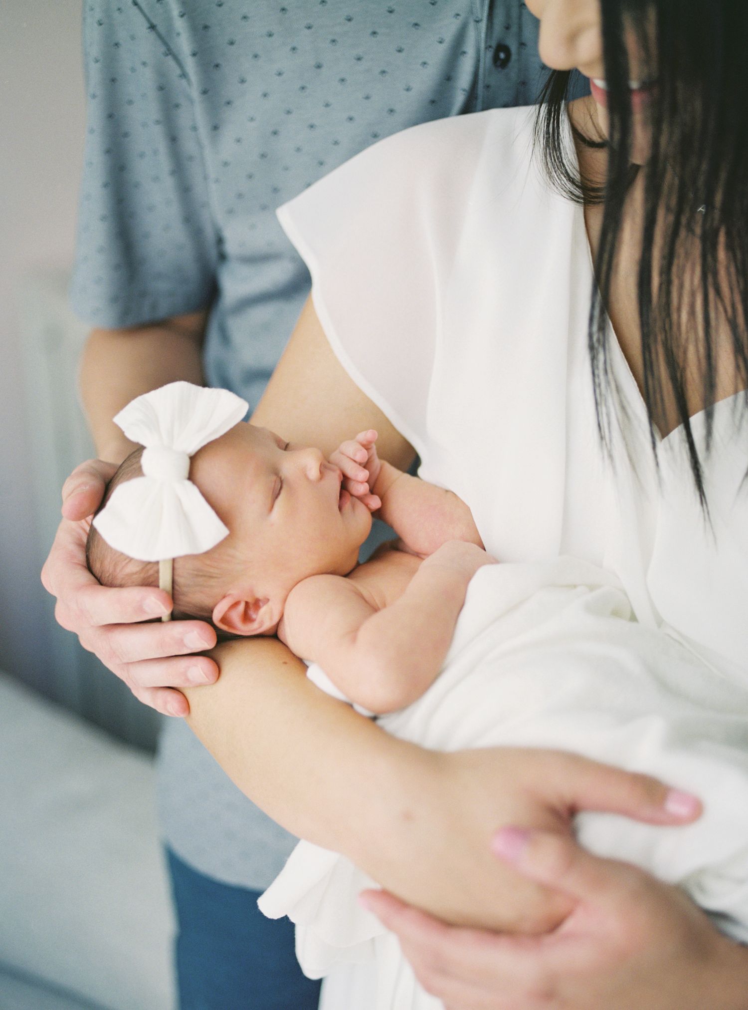Parents cradling sleeping newborn baby girl wearing white bow headband in soft natural light.