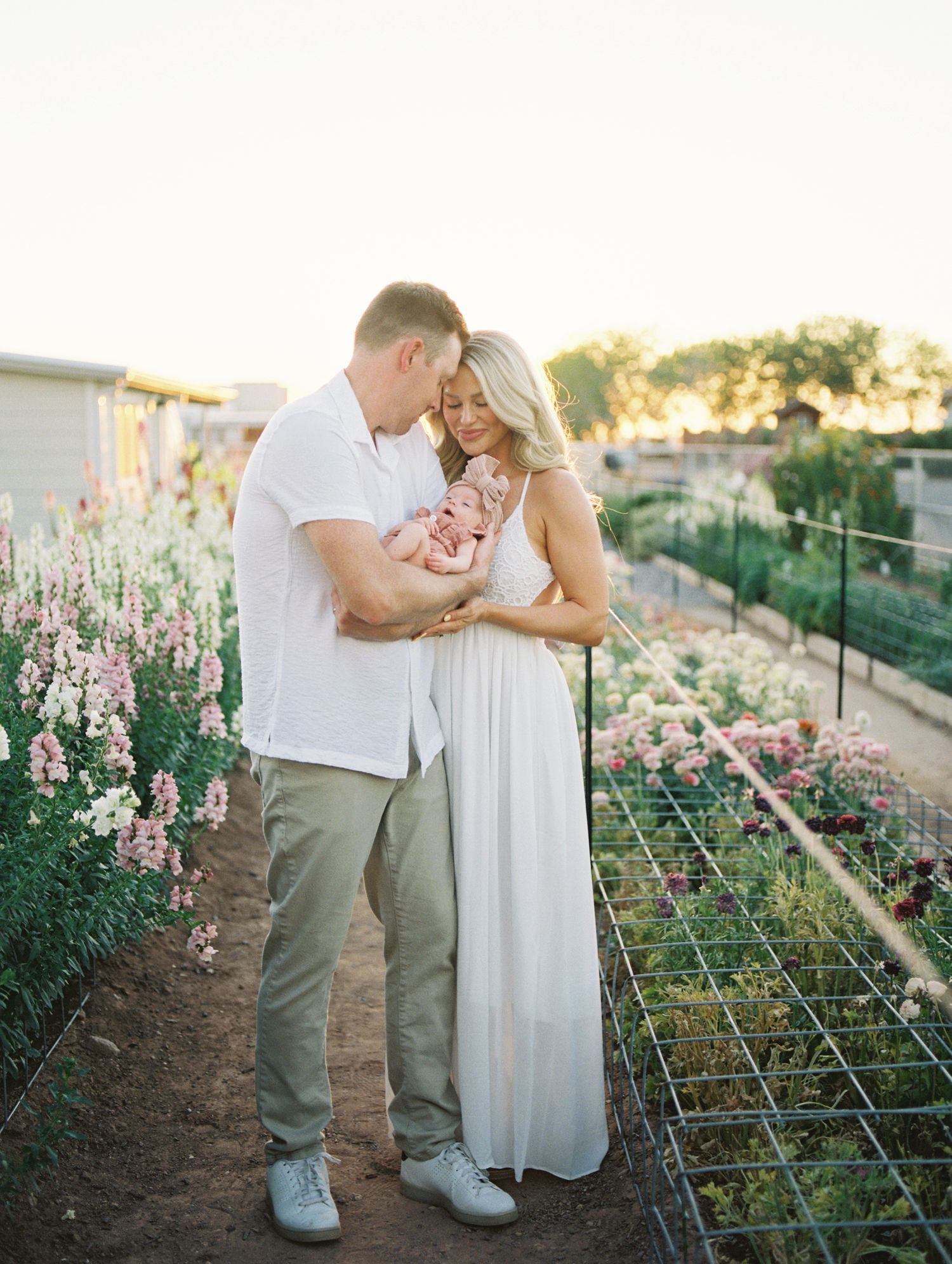 A couple in white clothing holds their newborn baby among colorful flower rows at golden hour.
