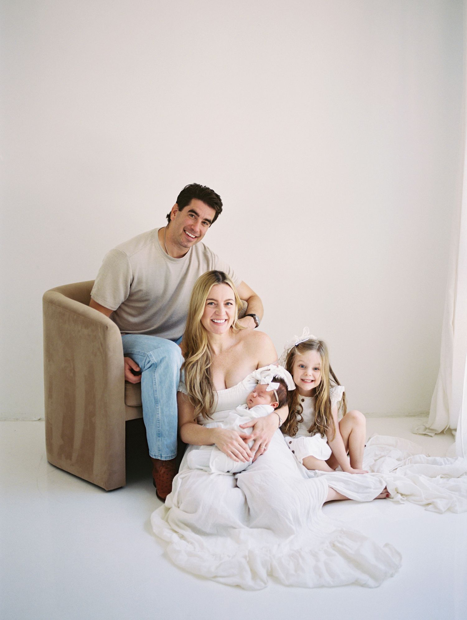 Happy family of four posing in bright white studio with newborn baby and smiling young daughter.