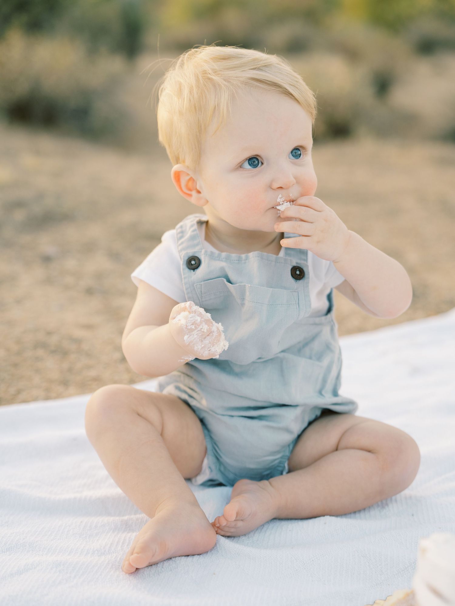 Cute baby boy in blue overalls eating birthday cake frosting while sitting on white blanket outdoors.