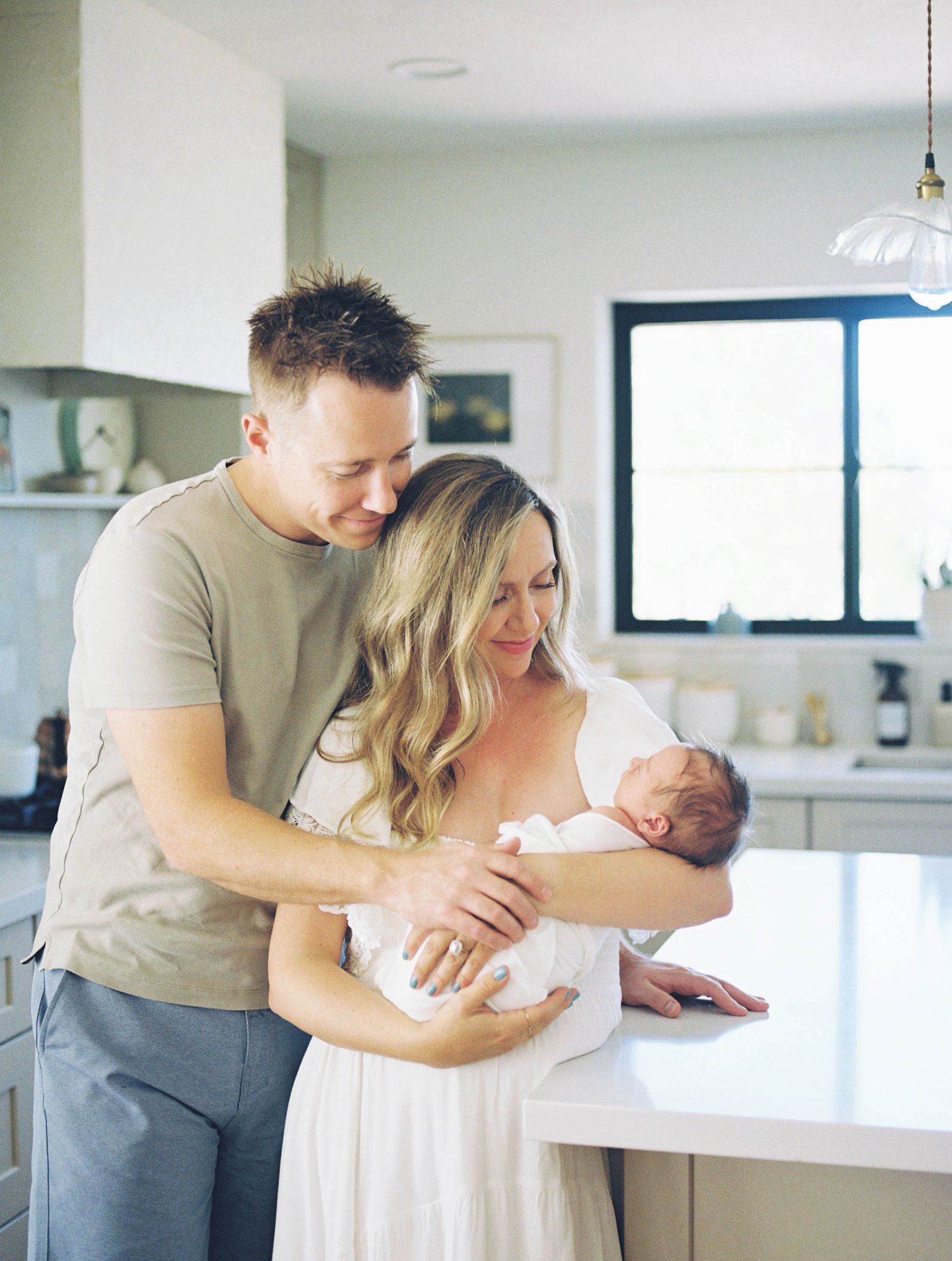 New parents in bright kitchen lovingly gazing at sleeping newborn baby, mother in white dress.