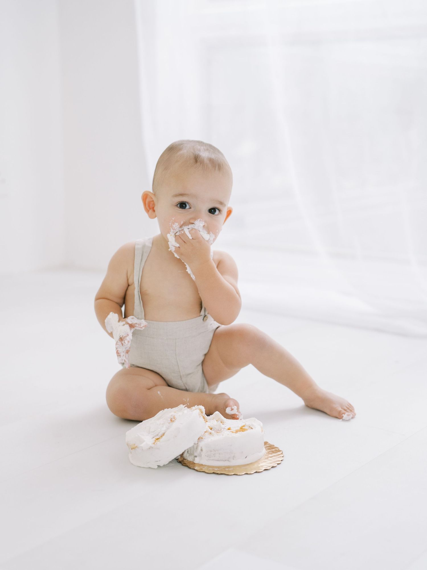 Adorable baby in gray romper smashing first birthday cake with frosting on face in white studio.