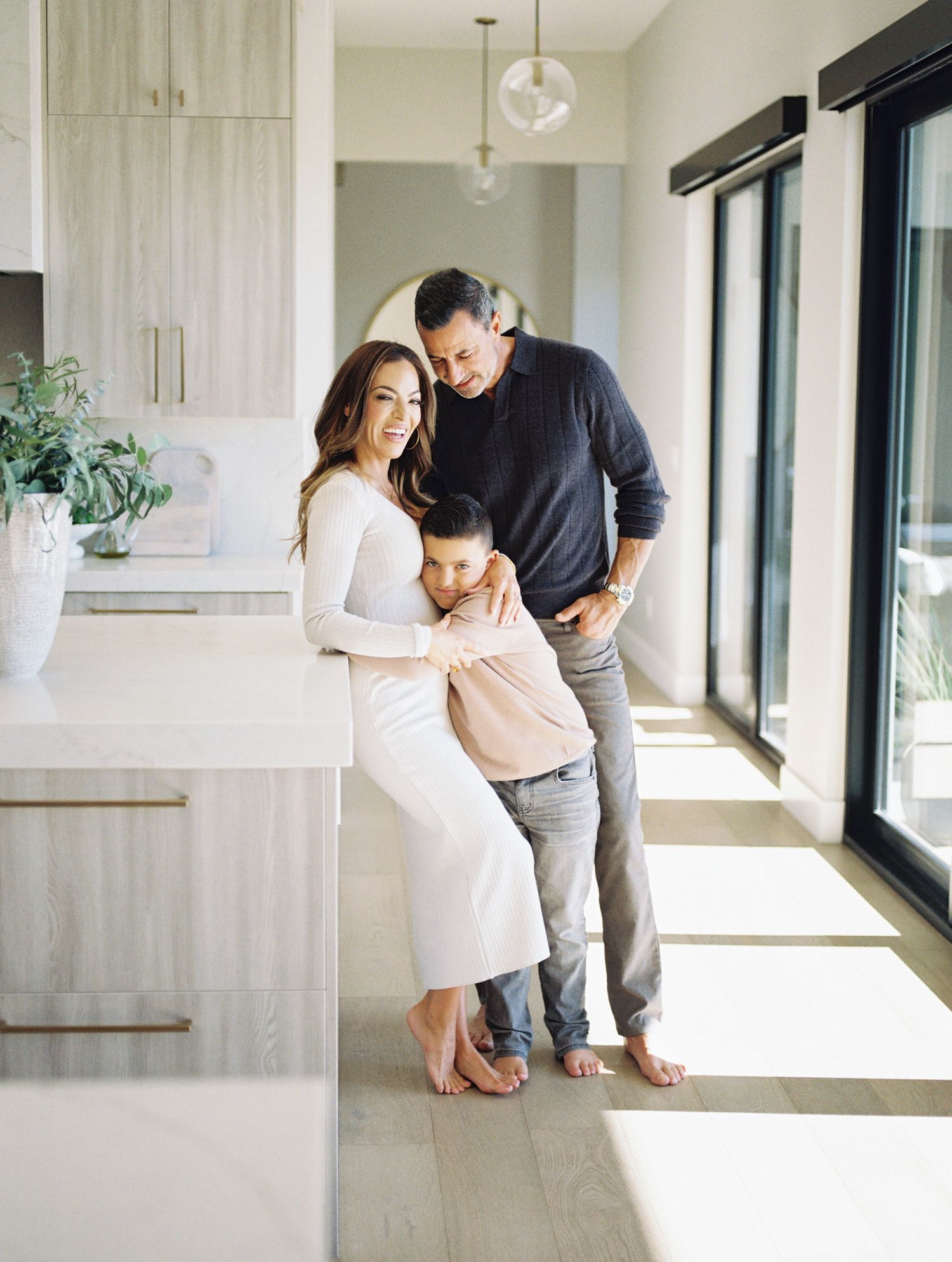 Pregnant woman, young son, and husband pose together in a bright modern kitchen with white countertops.