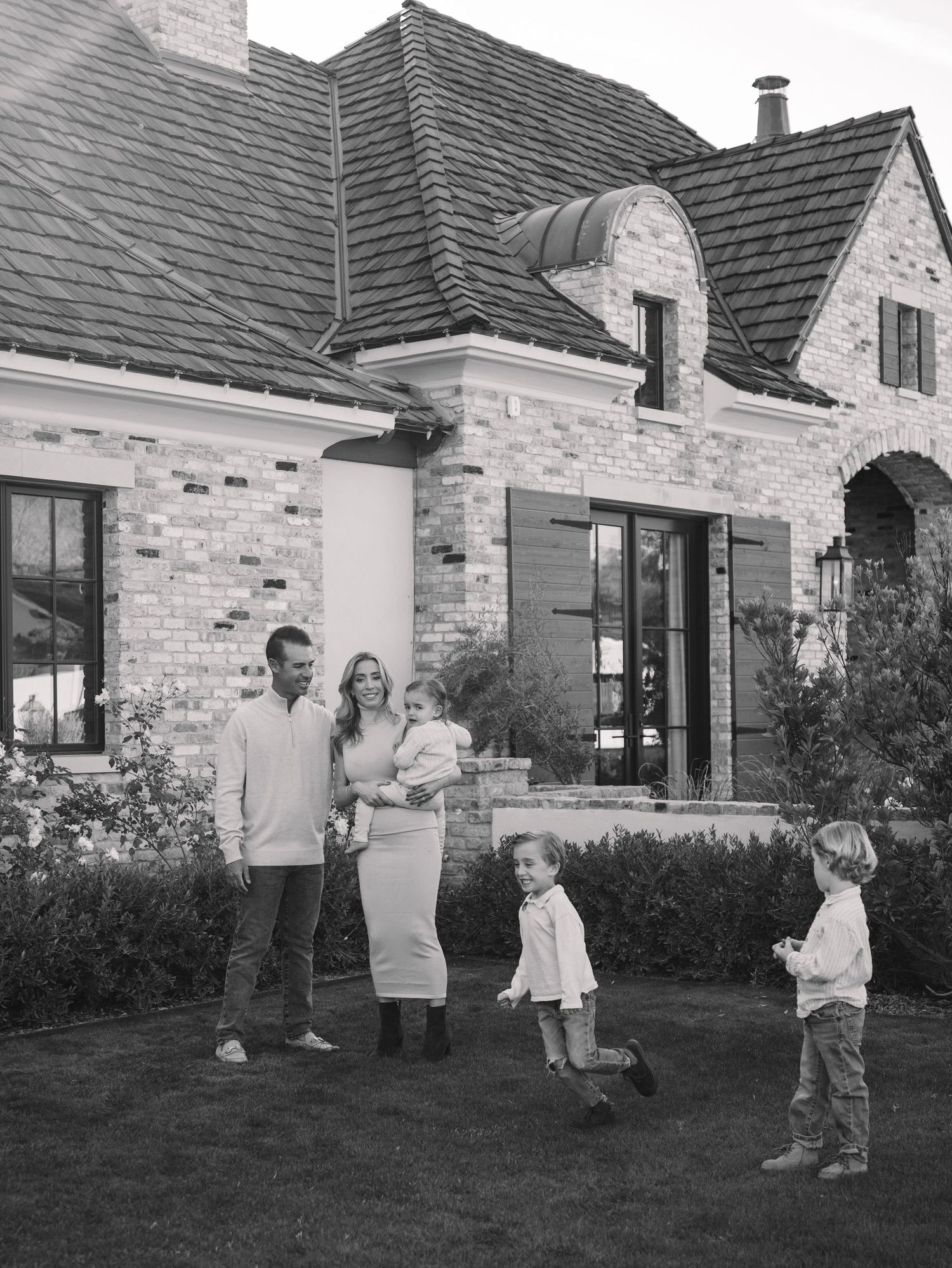 Black and white family portrait outside brick home with parents holding baby and two young boys playing on lawn.
