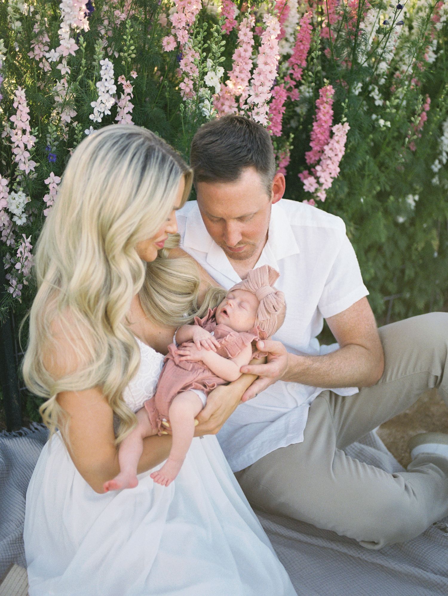 Couple in white attire tenderly holding newborn baby girl in pink outfit before floral backdrop.