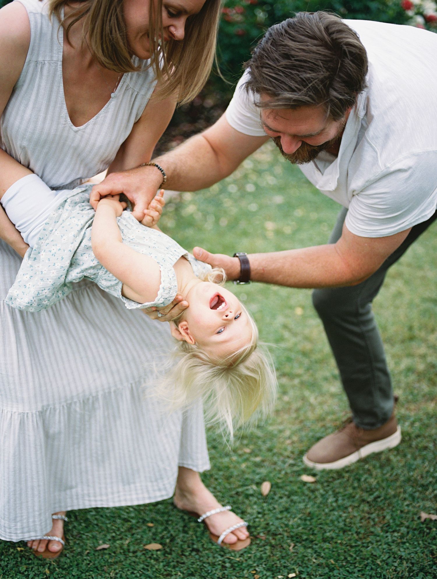 Joyful family moment as parents playfully flip their laughing blonde toddler upside down on green grass.