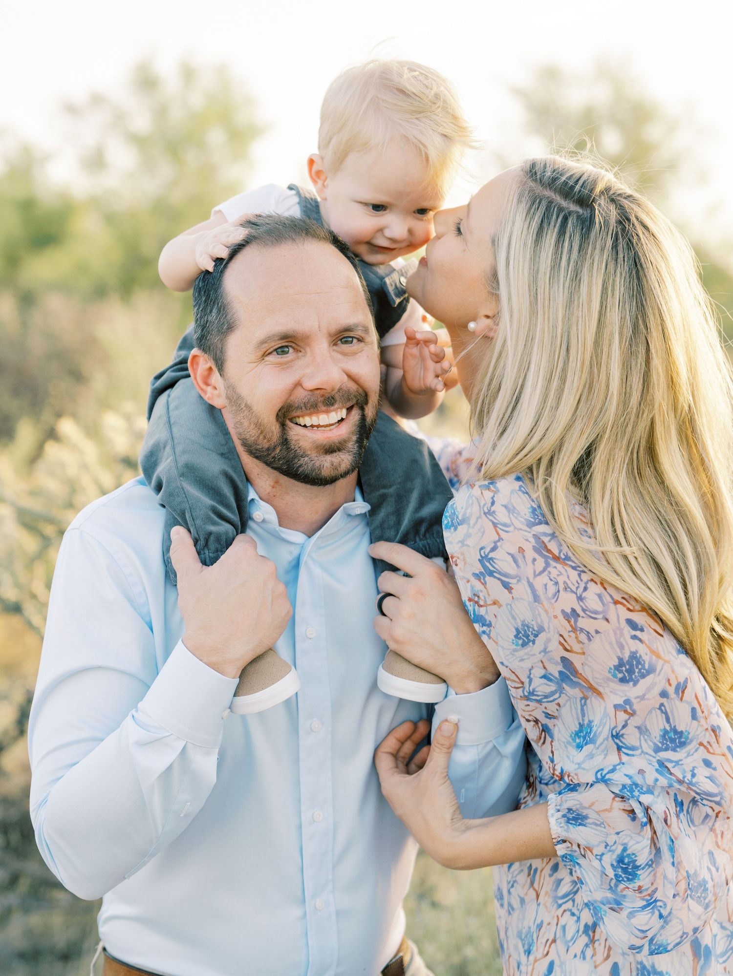 Happy family portrait with father carrying toddler on shoulders while mother kisses his cheek outdoors.