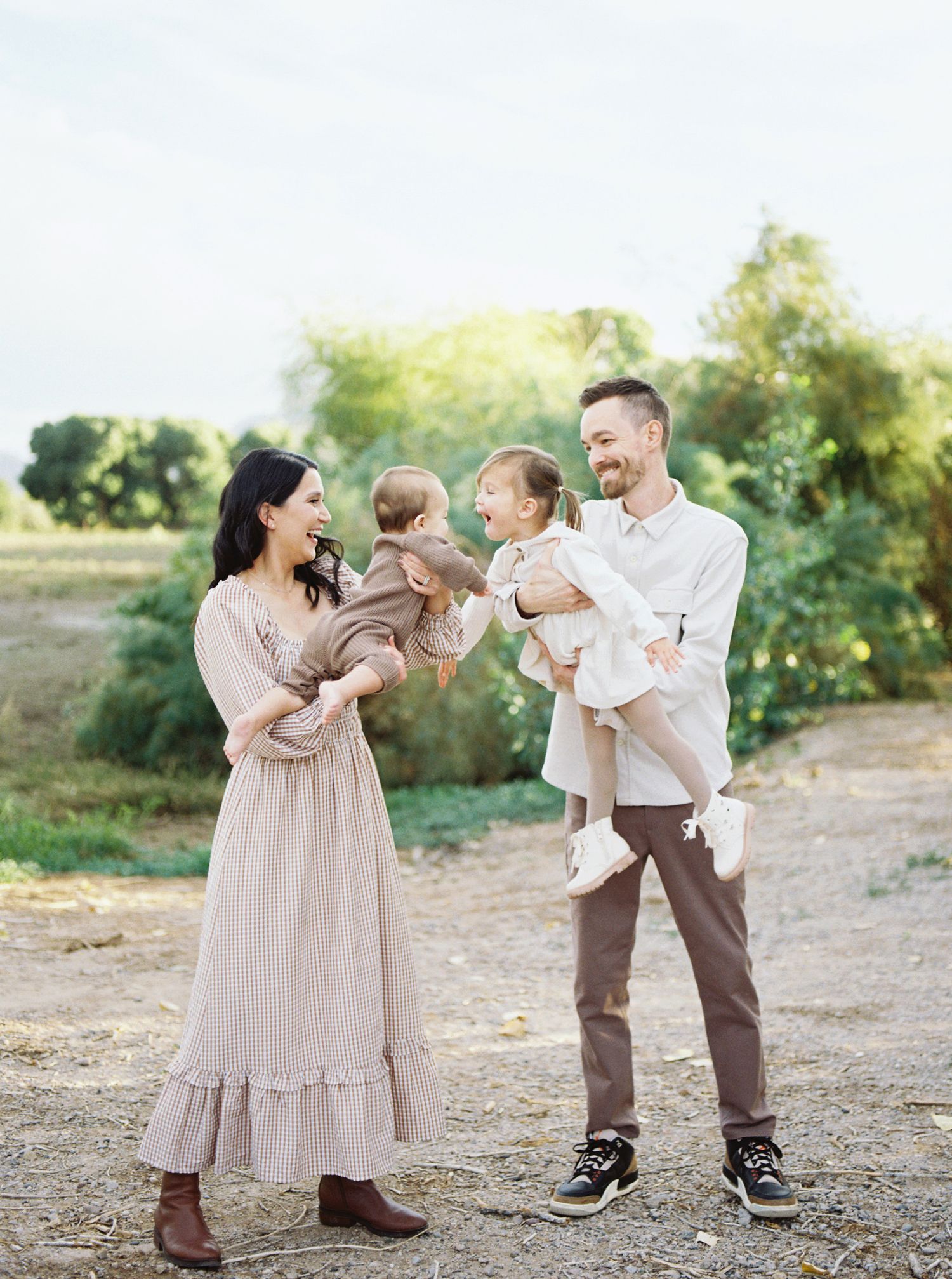 Happy family of four with two babies laughing together in sunny outdoor setting.