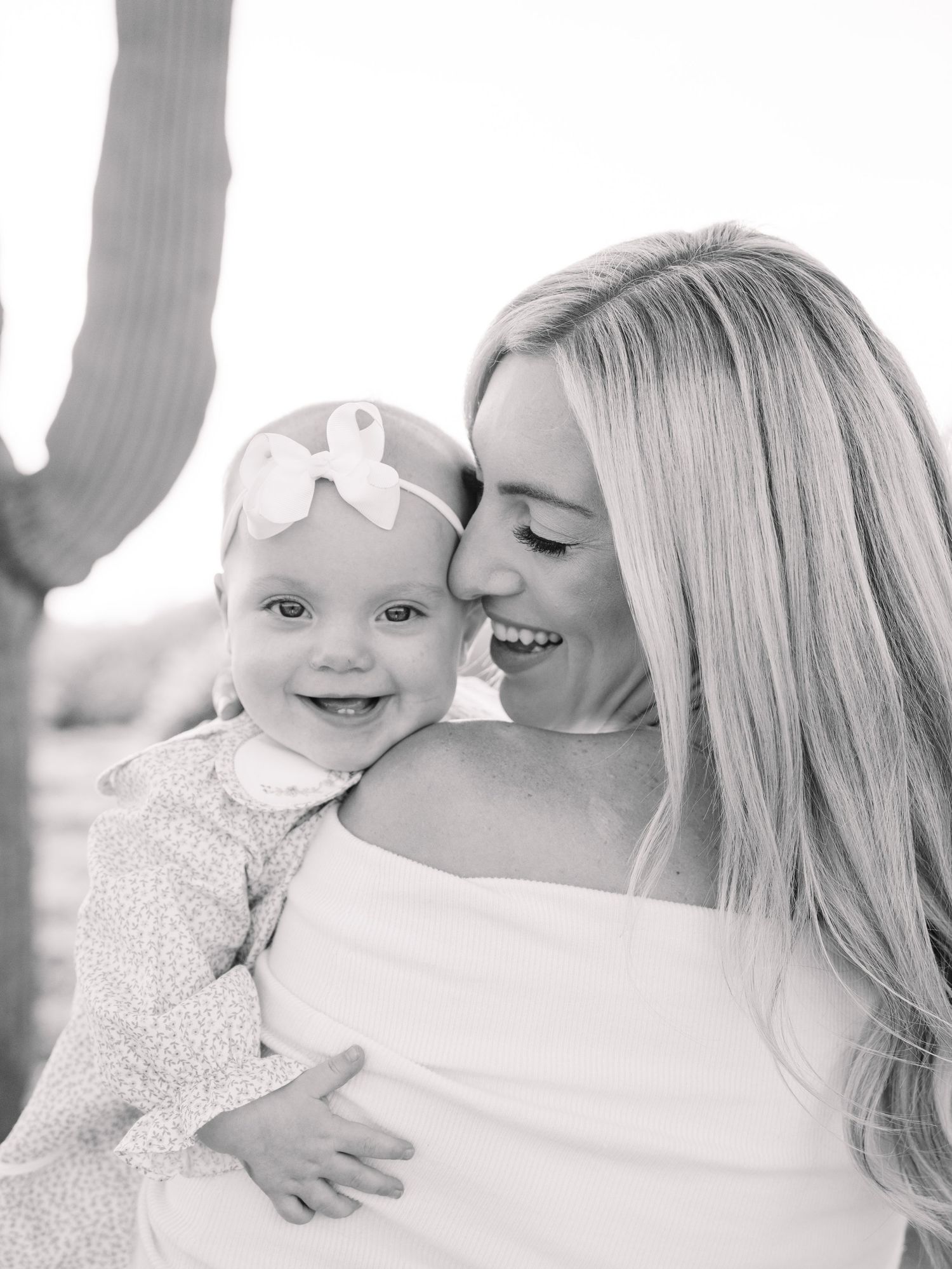 Black and white portrait of smiling blonde mother holding happy baby girl with bow headband.
