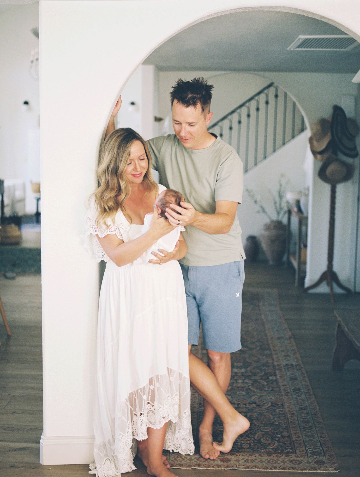 Couple in white home archway admiring newborn baby, mother in white lace dress, father in gray tee and shorts.