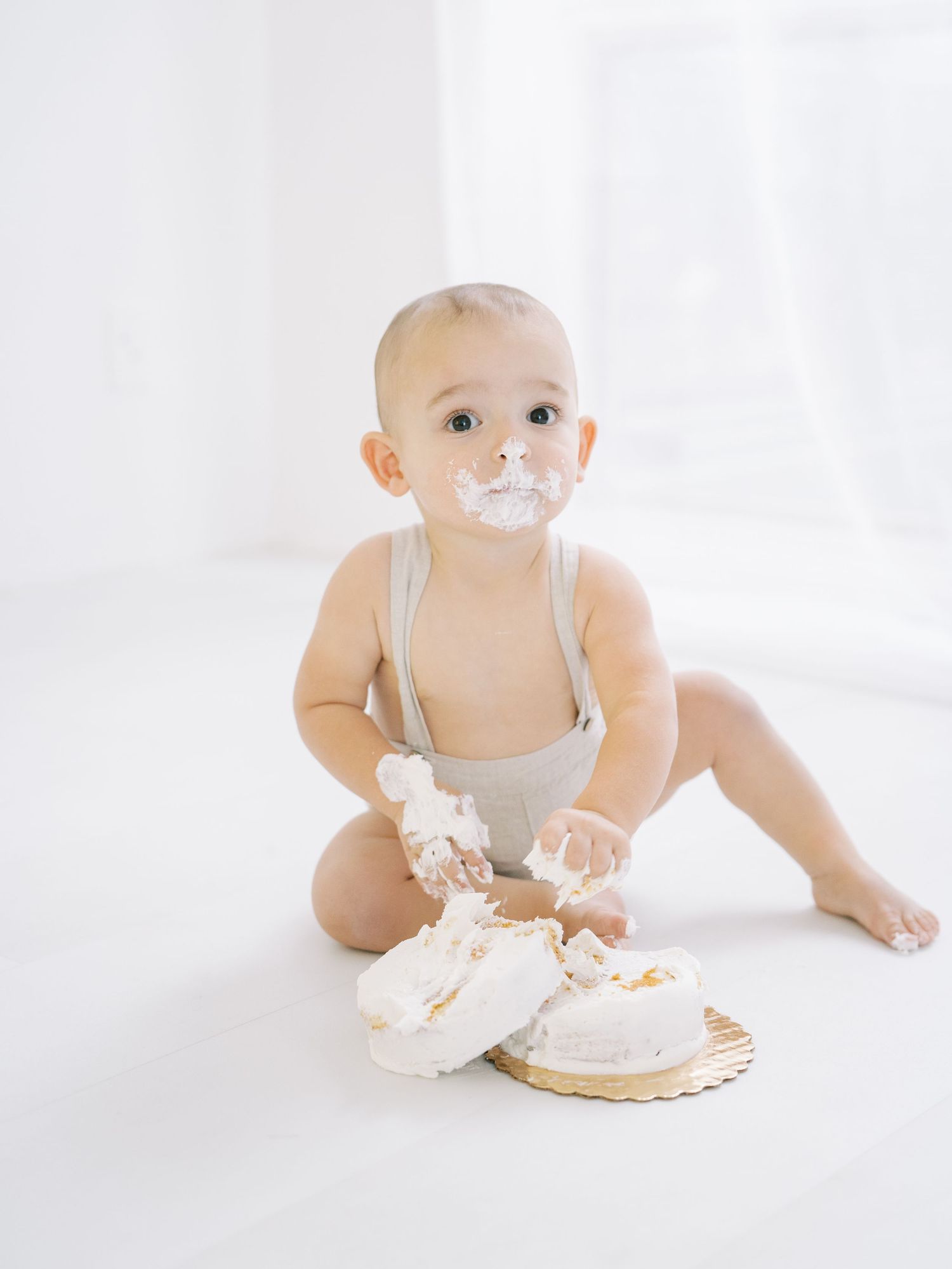 Adorable baby in linen romper with cake frosting on face during first birthday cake smash session.