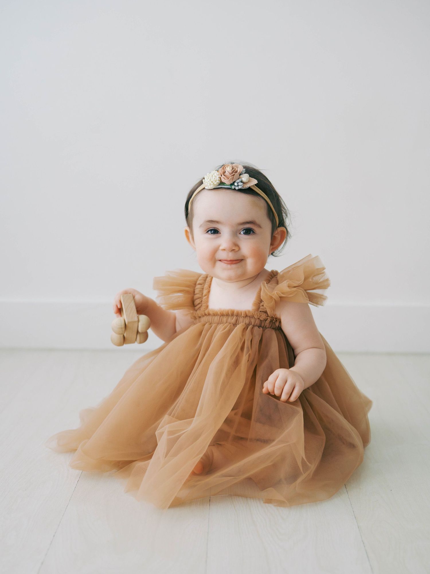 Beautiful baby girl in golden tulle dress with floral headband sitting on white floor smiling.