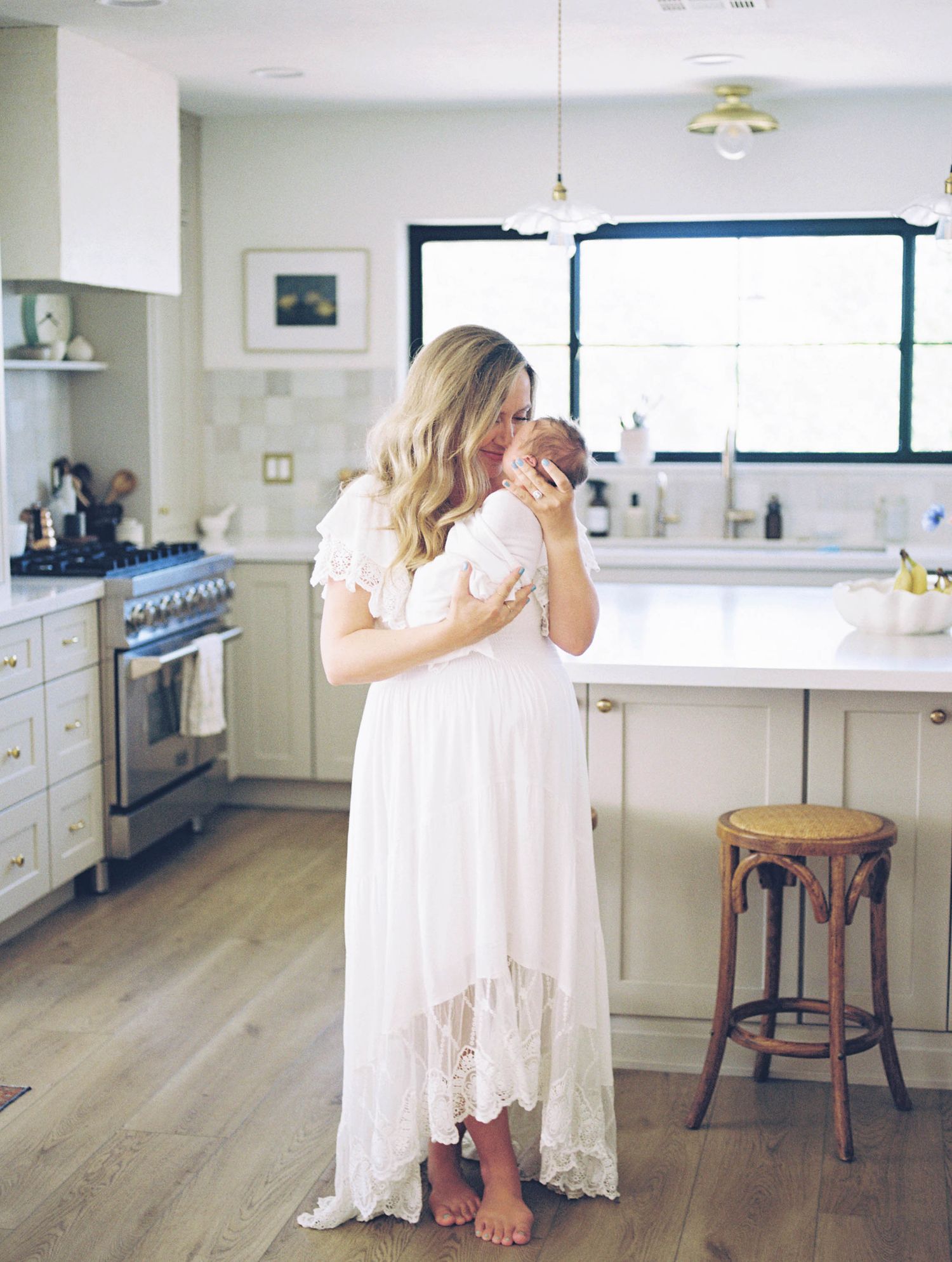 A woman in a white lace dress holds a newborn baby in a bright modern kitchen with hardwood floors.