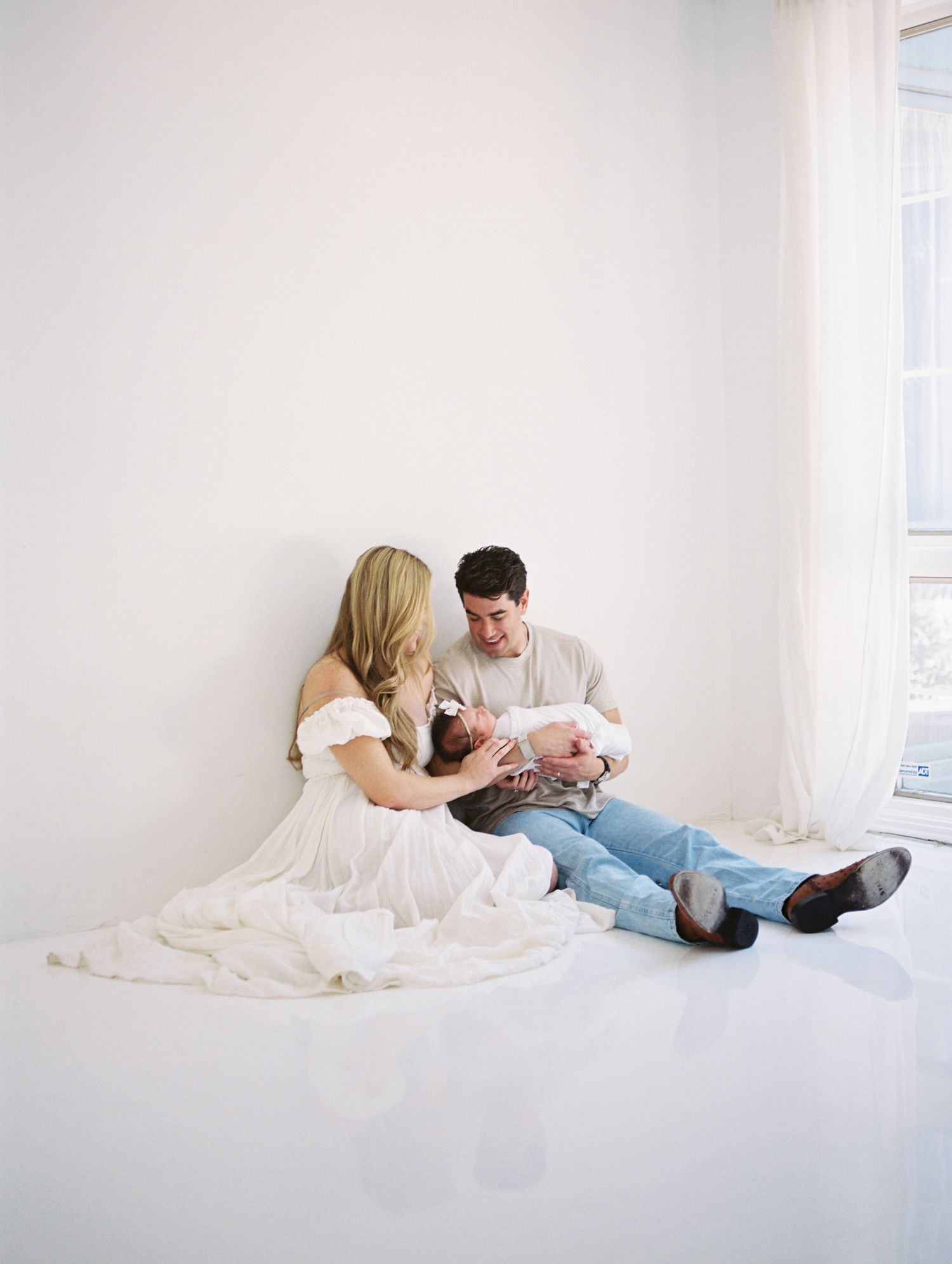 Parents sitting on white studio floor gazing lovingly at their newborn baby by a bright window.