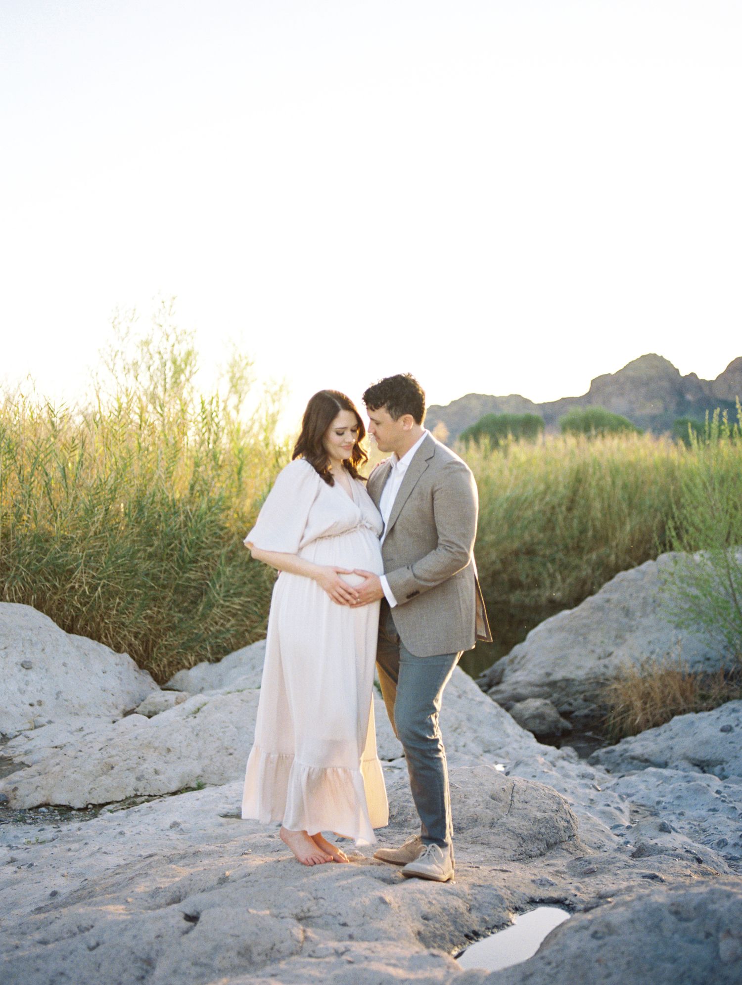 A pregnant couple shares a tender moment on rocky terrain with golden sunset light and lush greenery behind them.