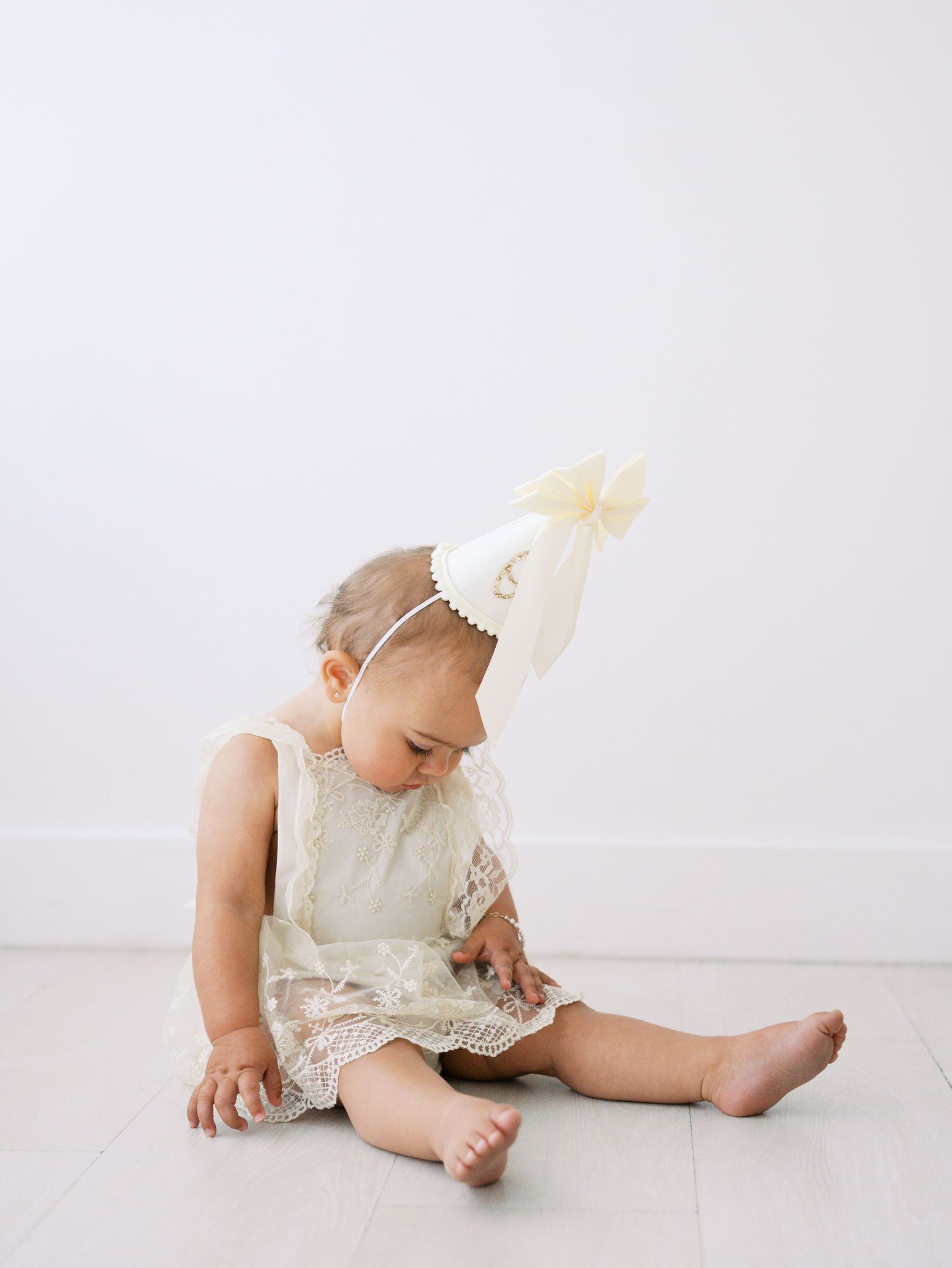 Baby girl in lace dress wearing party hat sitting on white floor looking down at hands.
