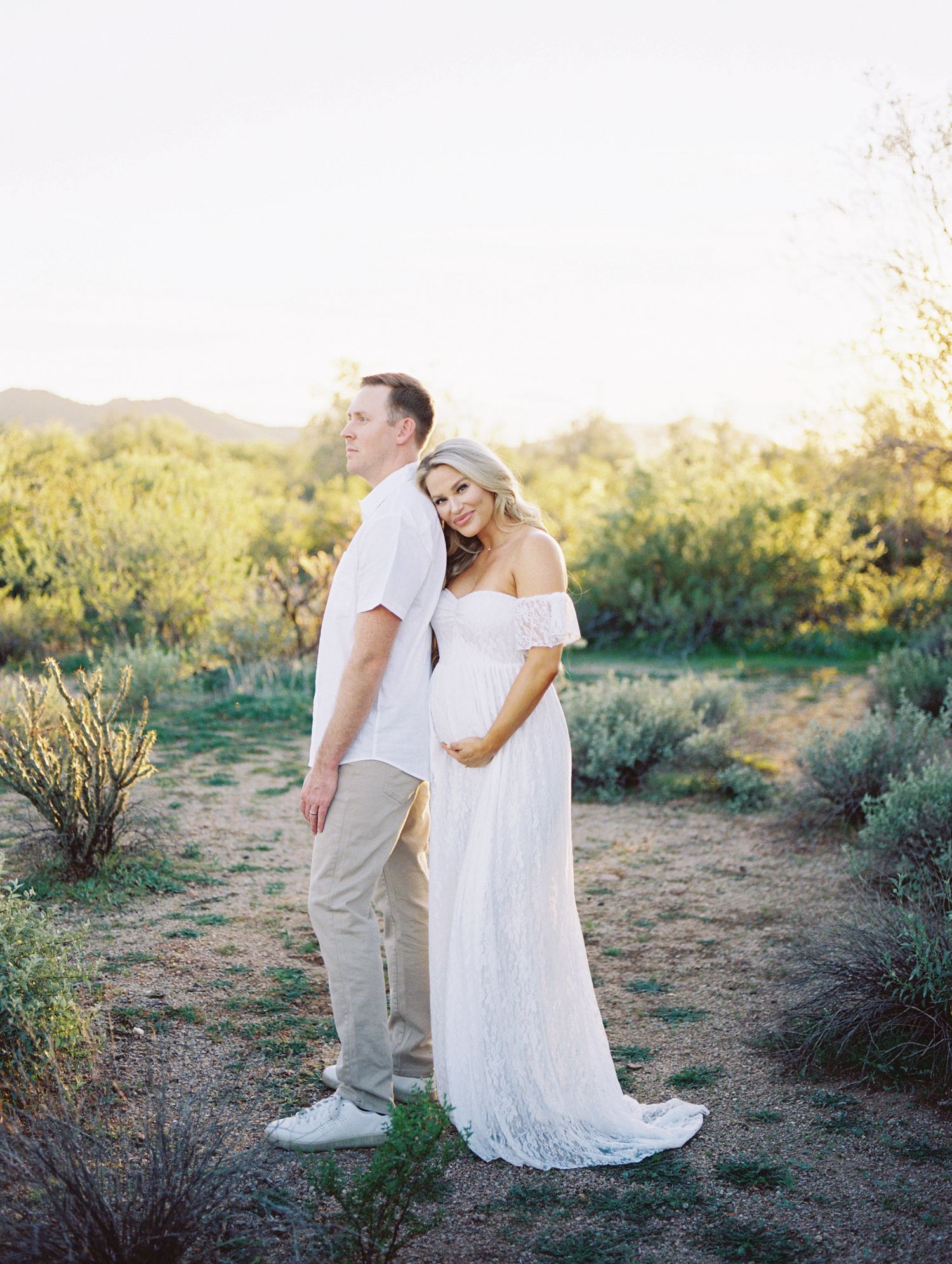 Expecting couple posing back-to-back in desert landscape at sunset, woman in white lace gown.