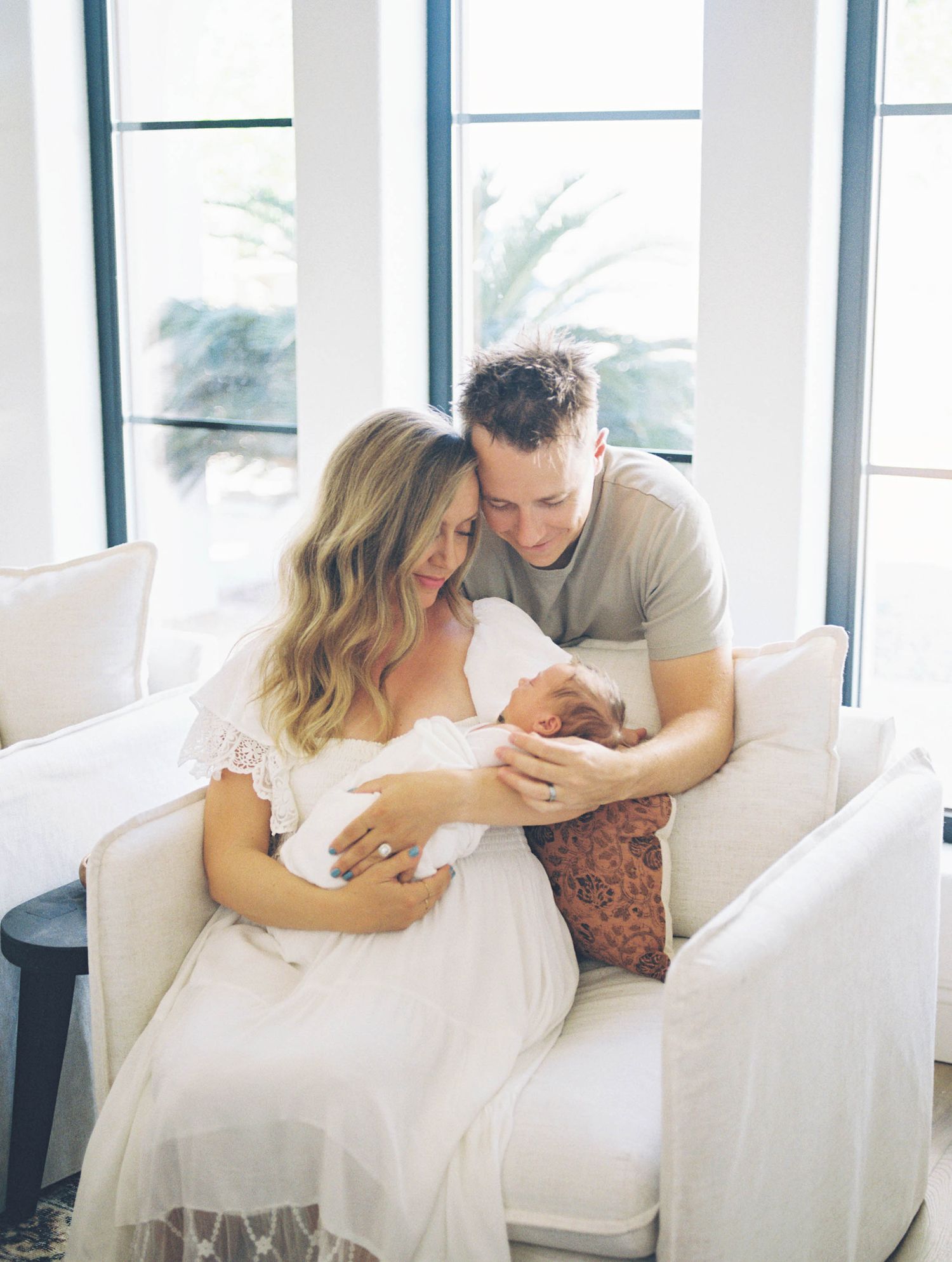 New parents sitting on white sofa gazing lovingly at newborn baby by window.
