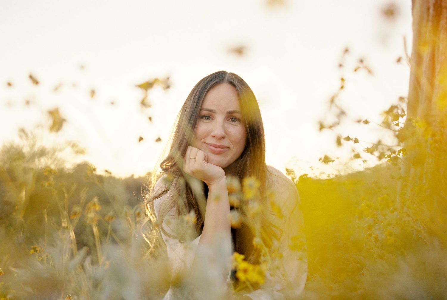 Young woman resting chin on hand surrounded by golden wildflowers in soft sunlight.