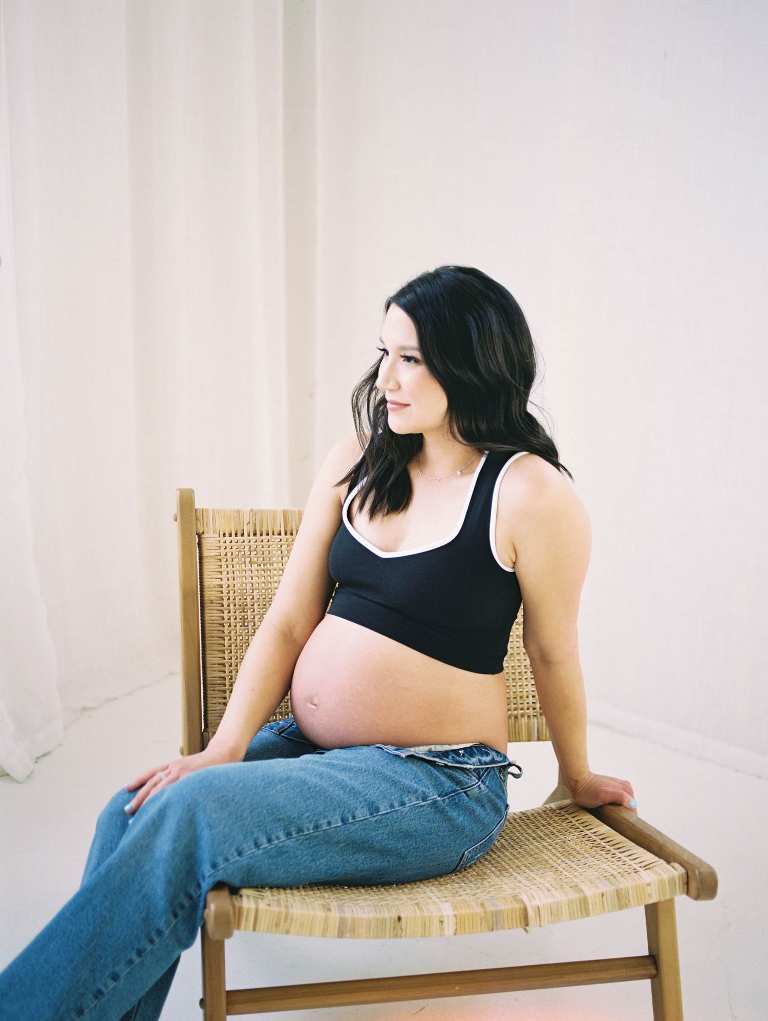 Pregnant woman in black crop top and jeans sitting in wicker chair during maternity session.