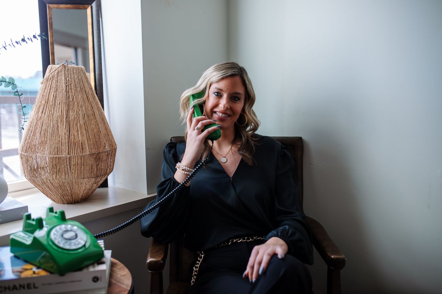 Smiling woman in a black blouse sits in a chair holding a green retro rotary phone receiver to her ear beside a woven lamp.