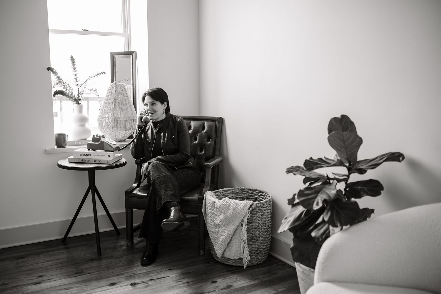 Woman sits in a tufted leather chair talking on the phone in a bright room with a side table, fiddle leaf fig plant and woven basket.