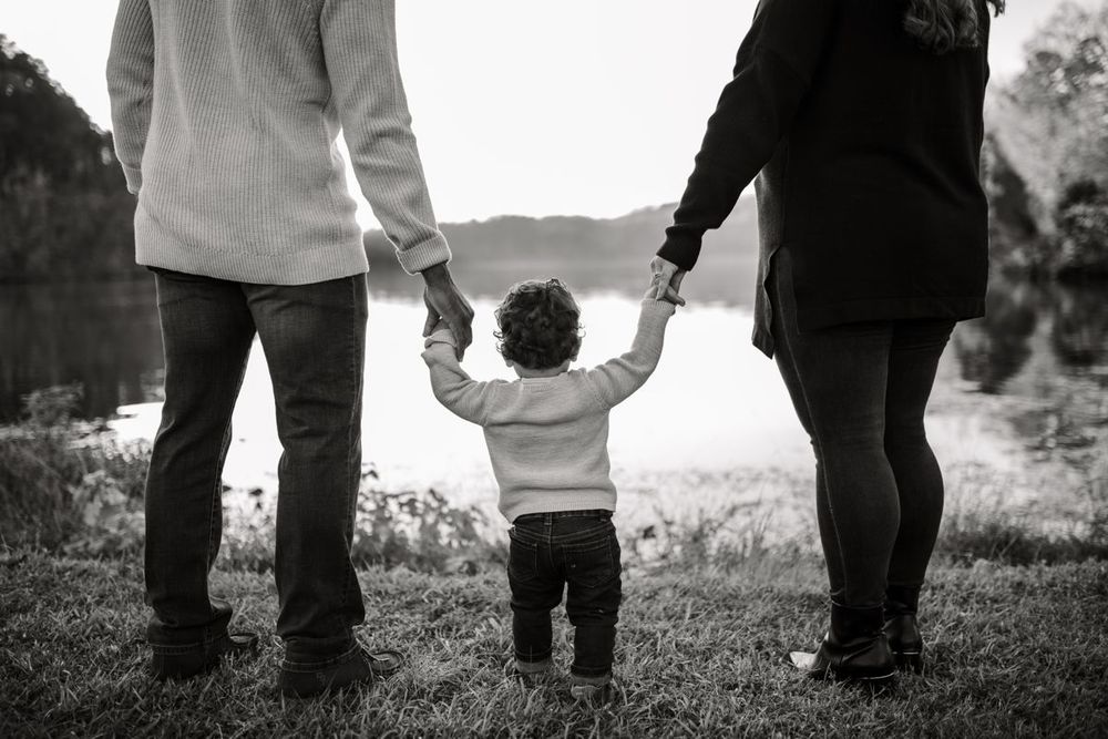 Black-and-white photo of a toddler holding both parents' hands by a lake