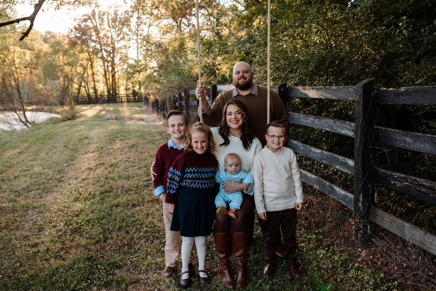 Family of six posing outdoors beside a wooden fence, with a man standing behind a woman holding a baby and three children in autumn light.