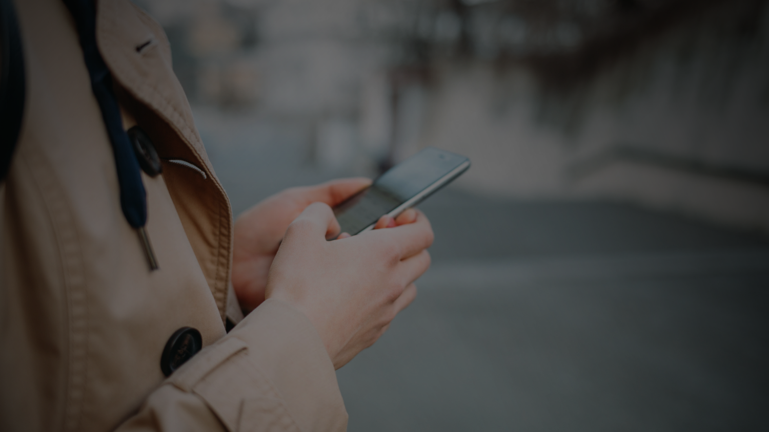 Person in a tan trench coat holds and texts on a smartphone while standing outdoors on a blurred city street.