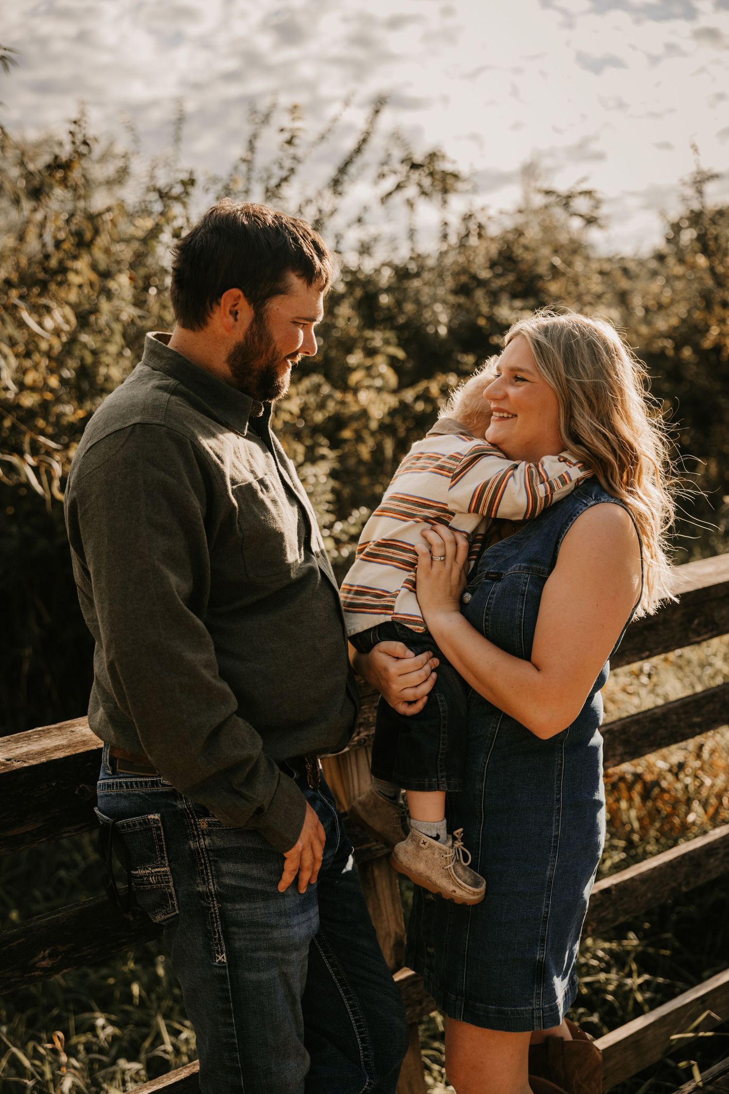 A bearded dad looks lovingly at his wife and toddler son as the mother laughs while holding the child near a rustic fence at sunset.