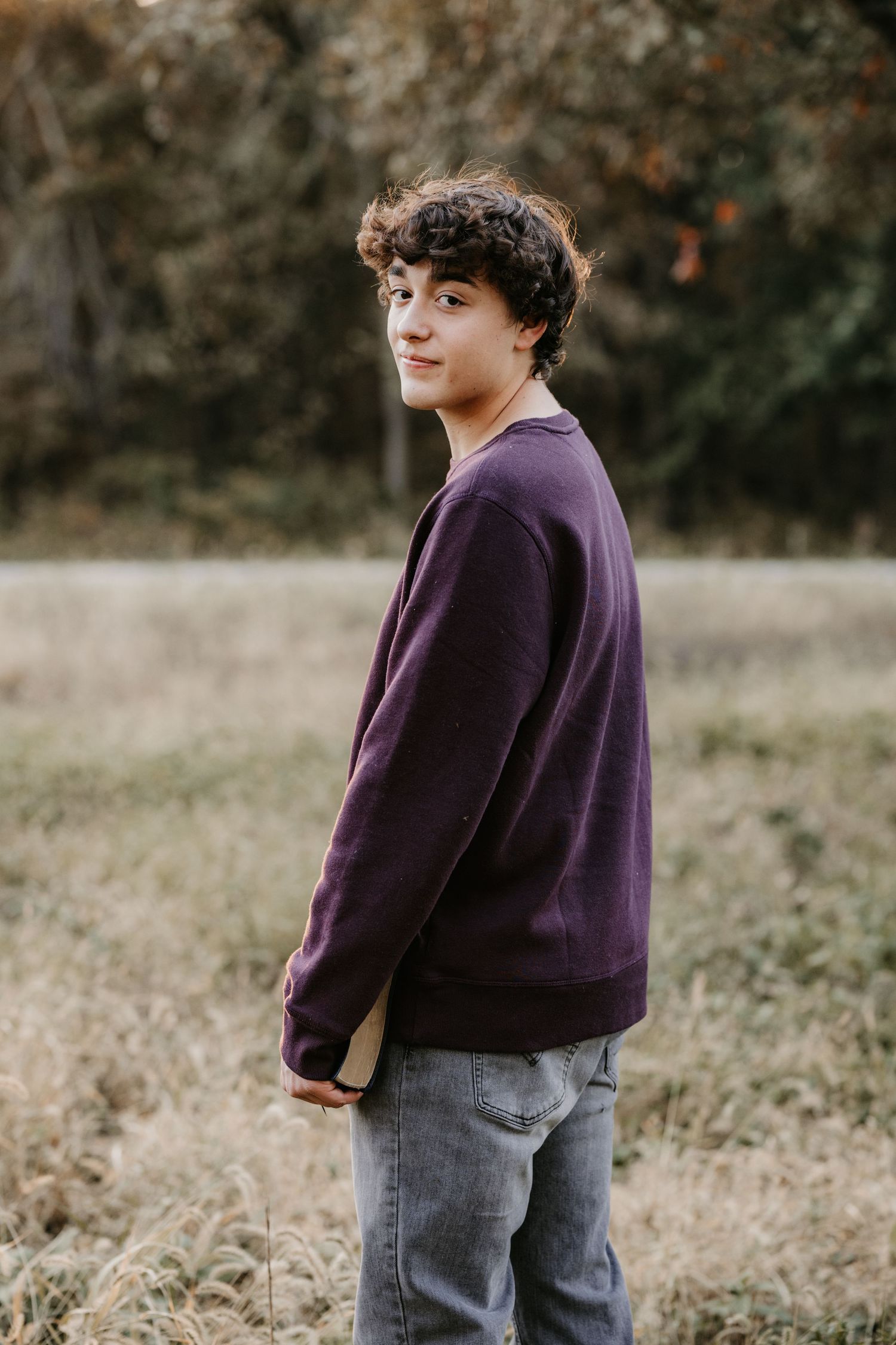 Young man in a maroon sweatshirt and gray jeans glances over his shoulder in a grassy field with a softly blurred forest behind him.