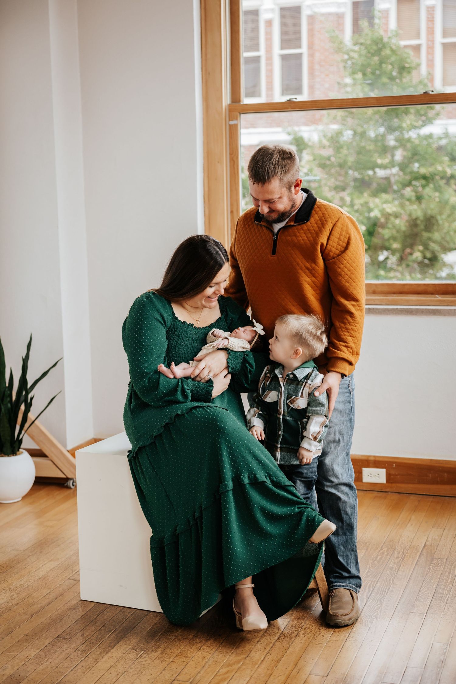 Mother in a green dress holds her newborn while dad and toddler son stand close in a bright studio family photo.