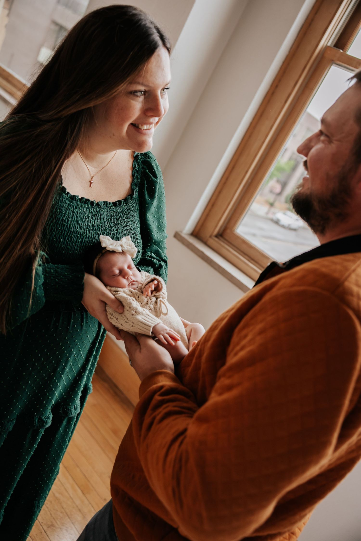 Mother in a green dress smiles down at her newborn baby as the father in an orange sweater looks on beside a sunny window.